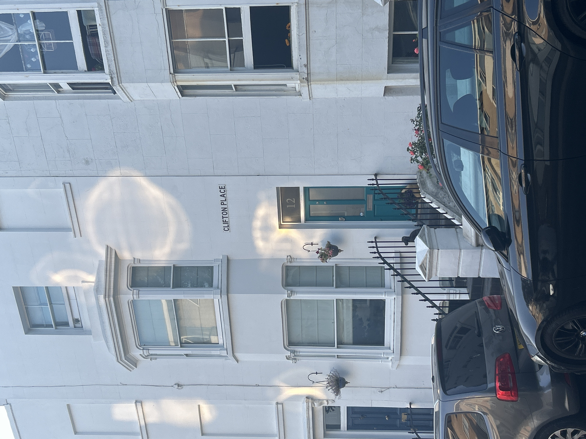 This image depicts the exterior of a white building in Clifton Place, Brighton, United Kingdom. The building has a white facade with several windows, a bay window, and a teal-colored door with the number 12 above it. The name "CLIFTON PLACE" is visible on the facade. Two cars, a dark gray Volkswagen and a black car, are parked in front of the building. There are hanging baskets with flowers near the door and on the side of the building. The scene is illuminated by sunlight, creating reflections on the building's surface.