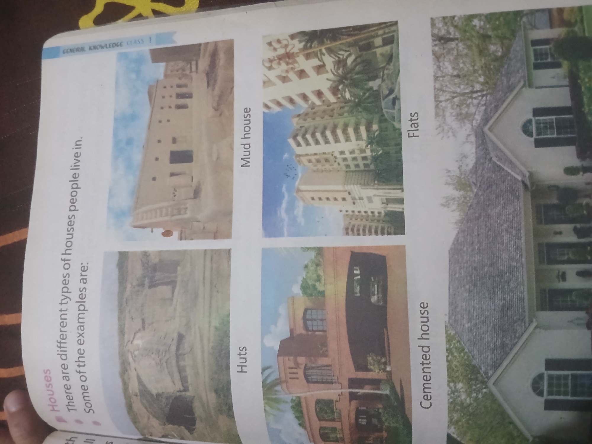 The image displays a page from a book titled "Houses." The text introduces the concept that there are different types of houses people live in and that the page will show examples. Below the introductory text, there are four distinct images of houses, each with a label.
The first image, labeled "Huts," shows several basic, low-lying structures with thatched roofs, set against a dry, earthy landscape. These appear to be simple dwellings, possibly in a rural or arid environment.
The second image, labeled "Mud house," depicts a long, flat-roofed building constructed from mud or adobe. It has several doorways and windows and appears to be a communal or multi-unit dwelling, with a light blue sky in the background.
The third image, labeled "Cemented house," features a multi-story residential building with a modern architectural design. It is constructed with concrete and has large windows and balconies, surrounded by greenery and palm trees. Cars are visible in the foreground, and the building's facade is light-colored.
The fourth image, labeled "Flats," shows a row of tall, apartment buildings, characterized by multiple stories and numerous windows. These buildings are also modern in design and appear to be located in an urban setting, with trees and a car visible in the foreground.
The image is a scanned page, showing a portion of the book's layout. The overall context is educational, providing visual examples of different housing types. There are no explicit indications or contextual clues within the image that would allow for a specific determination of its location in Peshawar City Tehsil, Pakistan. The provided images are generic representations of different housing styles.