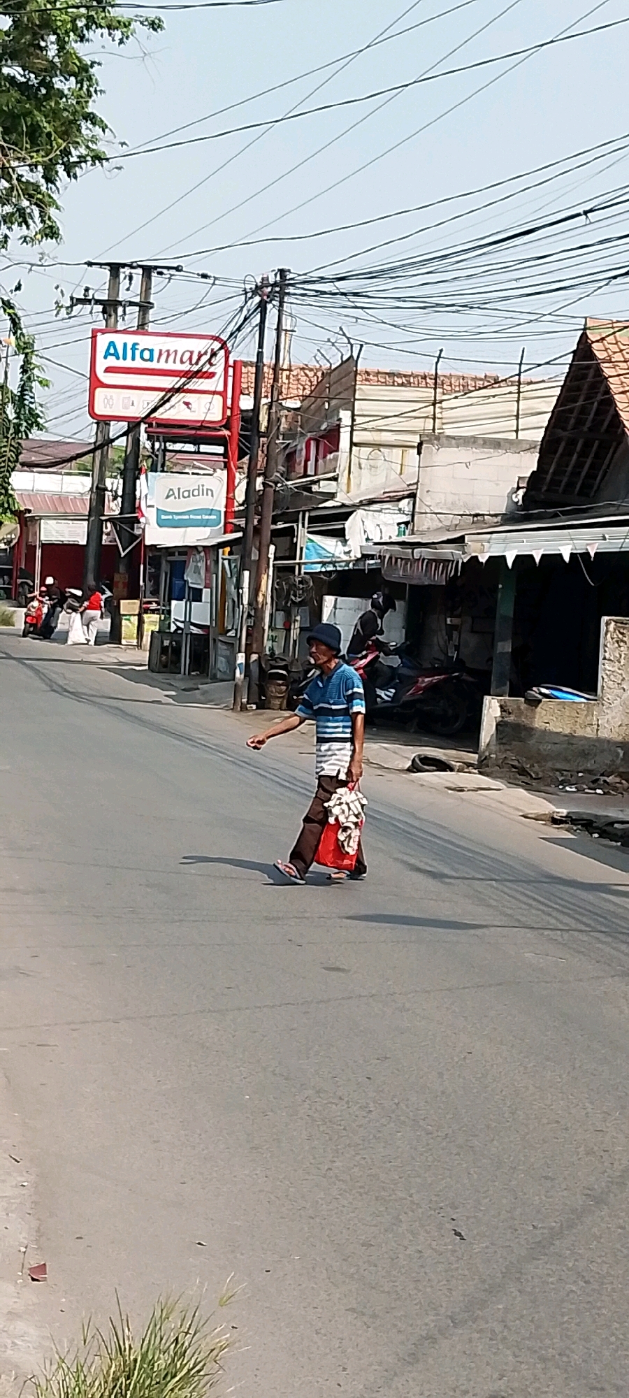 An elderly man wearing a blue and white striped shirt, brown pants, a blue hat, and flip-flops walks across a paved road in Babelan, Indonesia. He is carrying a red plastic bag filled with white items in his right hand, and his left arm is extended to the side. The road is in the foreground with visible lane markings. In the background, there are buildings, including a sign for "Alfamart" and another for "Aladin". There are also many electrical wires overhead. Other people and vehicles are visible in the background, including a motorcycle with two people on it. The scene appears to be a street in a commercial area.