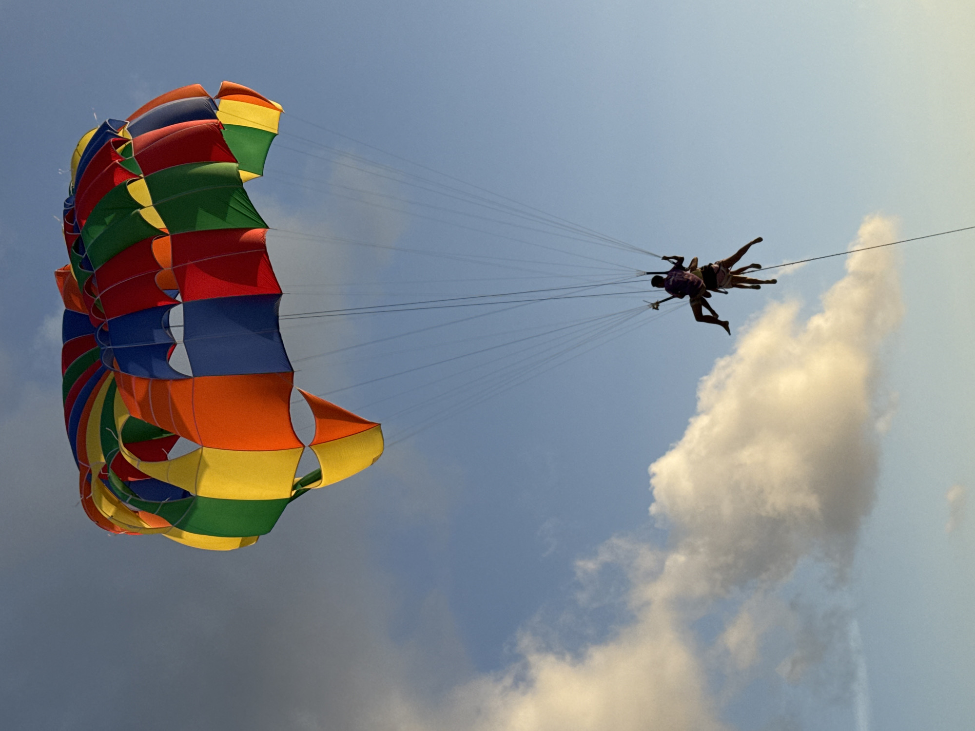 This is an image of two people parasailing over the ocean near Patong, Thailand. The sky is blue with some white clouds. The parasail is colorful, with sections of red, orange, yellow, green, and blue. The two individuals are suspended beneath the parasail by ropes, and they appear to be enjoying the ride. The ocean is not visible in the frame. The overall impression is one of a fun and adventurous activity.