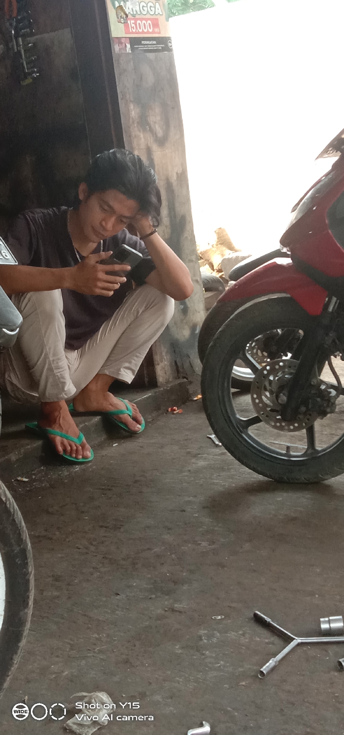 A young man in a black t-shirt and white pants crouches near a motorbike repair shop. He looks at his smartphone, resting his head on his left hand. He wears green flip-flops. A red motorcycle is next to him, with its front wheel clearly visible. A few tools lie on the concrete floor. A poster hangs on the wall behind him.