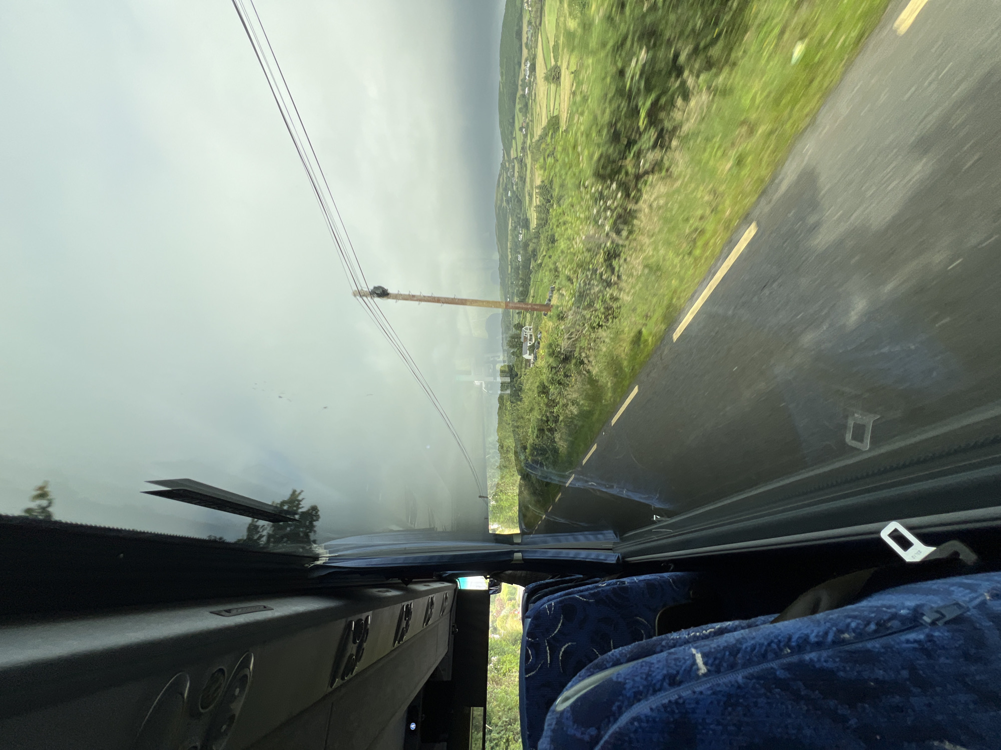 This is a photograph taken from inside a vehicle, possibly a bus, looking out the windshield. The image is oriented sideways, making it appear as if the vehicle is traveling vertically. Outside the windshield, a road curves to the right, bordered by a lush green grassy verge. In the distance, rolling hills covered in fields and dotted with houses suggest a rural landscape. The sky is overcast with a layer of dark clouds, indicating potentially inclement weather, but there are also patches of lighter sky, hinting at the possibility of the sun breaking through.
In the foreground, inside the vehicle, the dashboard and part of a seat are visible. The dashboard appears to be made of dark material, with some reflective surfaces. A blue, patterned seat is visible on the right side of the frame, partially covered by a dark blanket or fabric. A seatbelt buckle is also visible, suggesting that the occupants are secured.
A prominent feature in the middle ground is a wooden utility pole supporting overhead wires. The wires stretch across the frame, disappearing into the hazy sky. The overall impression is that of a journey through a scenic, green, and somewhat moody rural area. The time of day is not definitively clear due to the overcast conditions, but it appears to be daytime. The location is identified as Baltoney Upper, Ireland. There is no visible text in the image.