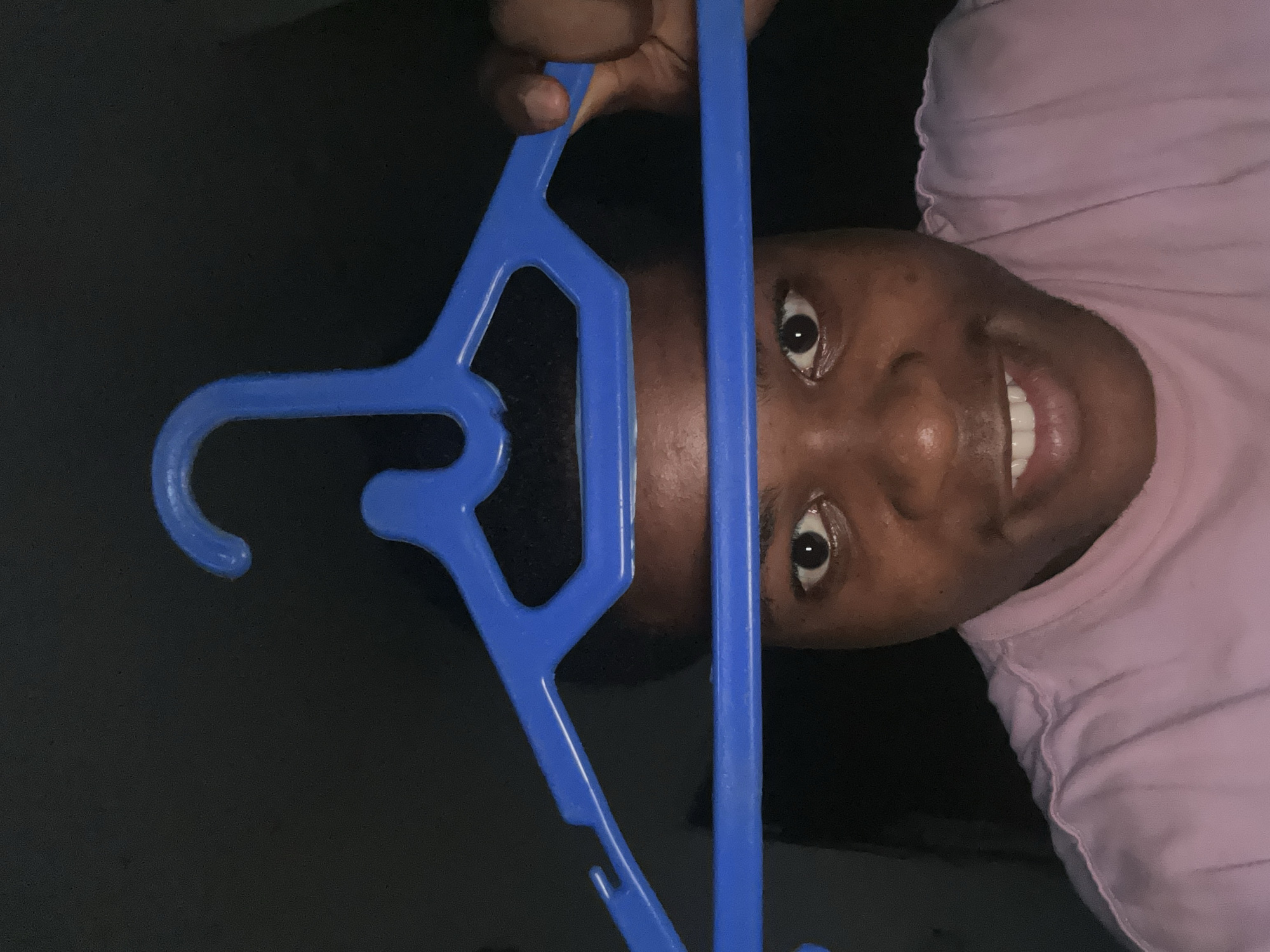 A person is holding a blue plastic hanger in front of their face, obscuring their forehead and hair. Their eyes are visible, looking directly at the camera. They are wearing a pink t-shirt. The background is dark and out of focus, suggesting an indoor setting. The image appears to be a selfie. The location is Ihiagwa, Nigeria.