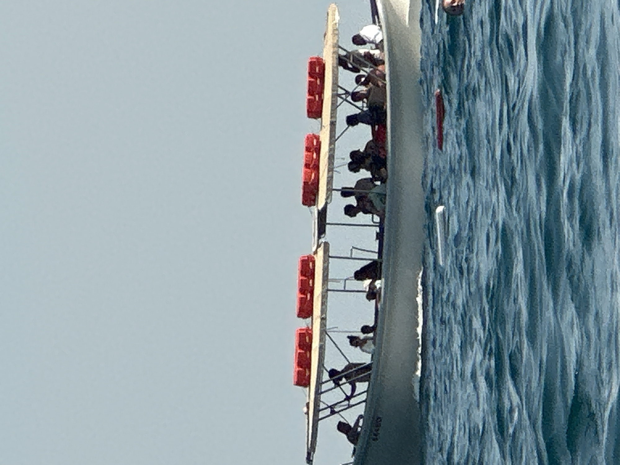 In Boreti, Montenegro, a white motorboat with a canopy is carrying approximately twelve individuals. The boat is navigating through moderately choppy blue water under a clear sky. Two bright red cylindrical objects, possibly life preservers, are secured to the top of the boat's canopy. The boat has a visible serial number on its side, "84490". In the foreground, a person's head is visible above the water on the right side of the image, suggesting they may be swimming or in distress. Several other red objects are floating in the water, some appearing to be buoys or debris. The overall scene depicts a daytime activity on the water.