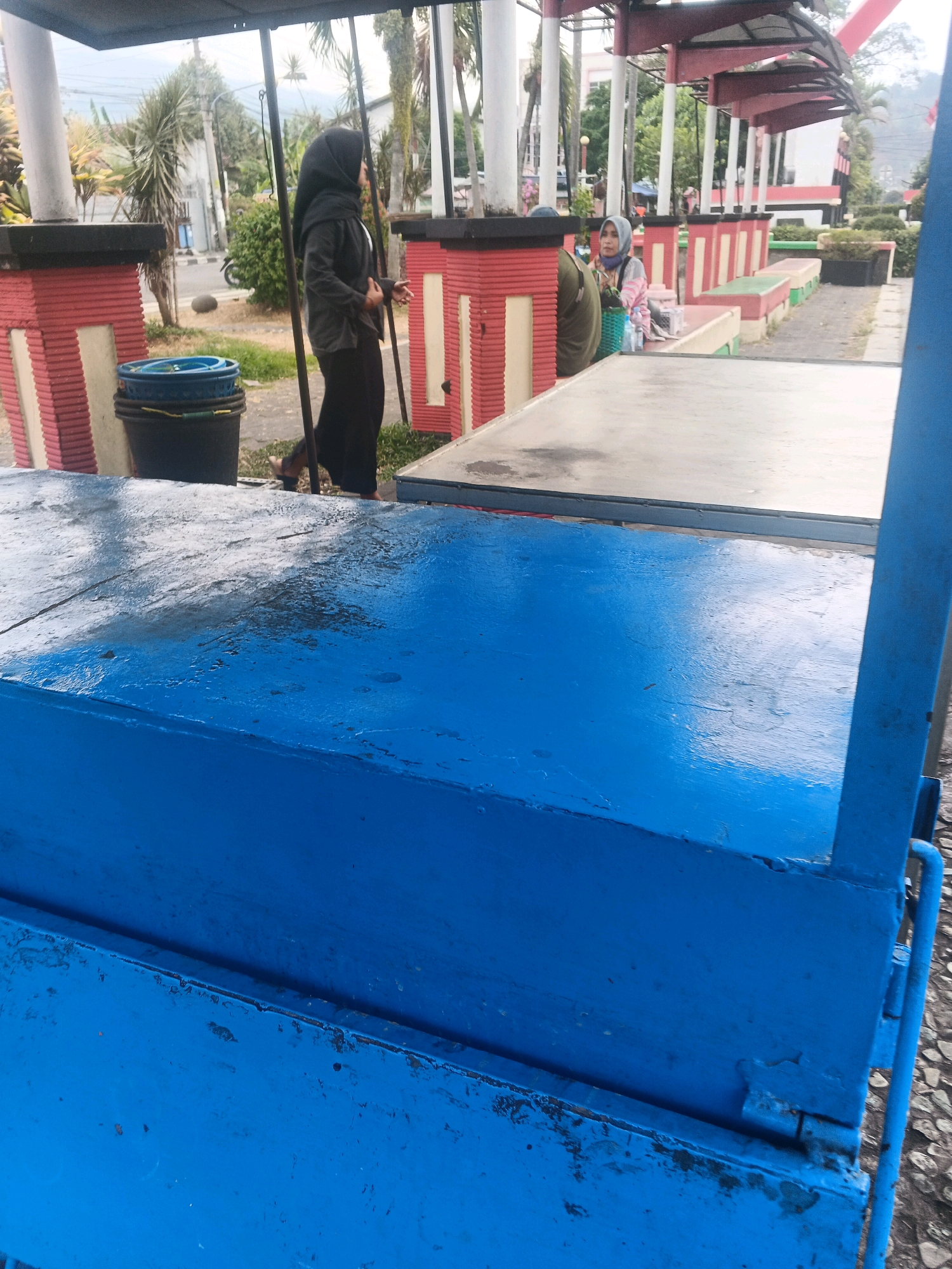In Ungaran, Indonesia, a scene unfolds in an outdoor area with paved ground and grassy patches, suggesting a public space such as a park or marketplace. In the foreground, a weathered blue wooden structure, possibly a vendor's cart or table, dominates the view. Its surface is worn and shows signs of use, with peeling paint and scuff marks.
In the mid-ground, a woman wearing a black hijab and long black clothing walks past a dark waste bin with a blue lid. To her right, a section of a covered seating area with red and white pillars and a red roof is visible. Further in the background, other individuals are present, one wearing a light-colored hijab and a pink and white patterned top, sitting on a bench. Behind them, a white building with a red trim can be seen, along with trees and vegetation, indicating an urban or semi-urban setting. The overall atmosphere appears to be one of daily activity in a local public space.