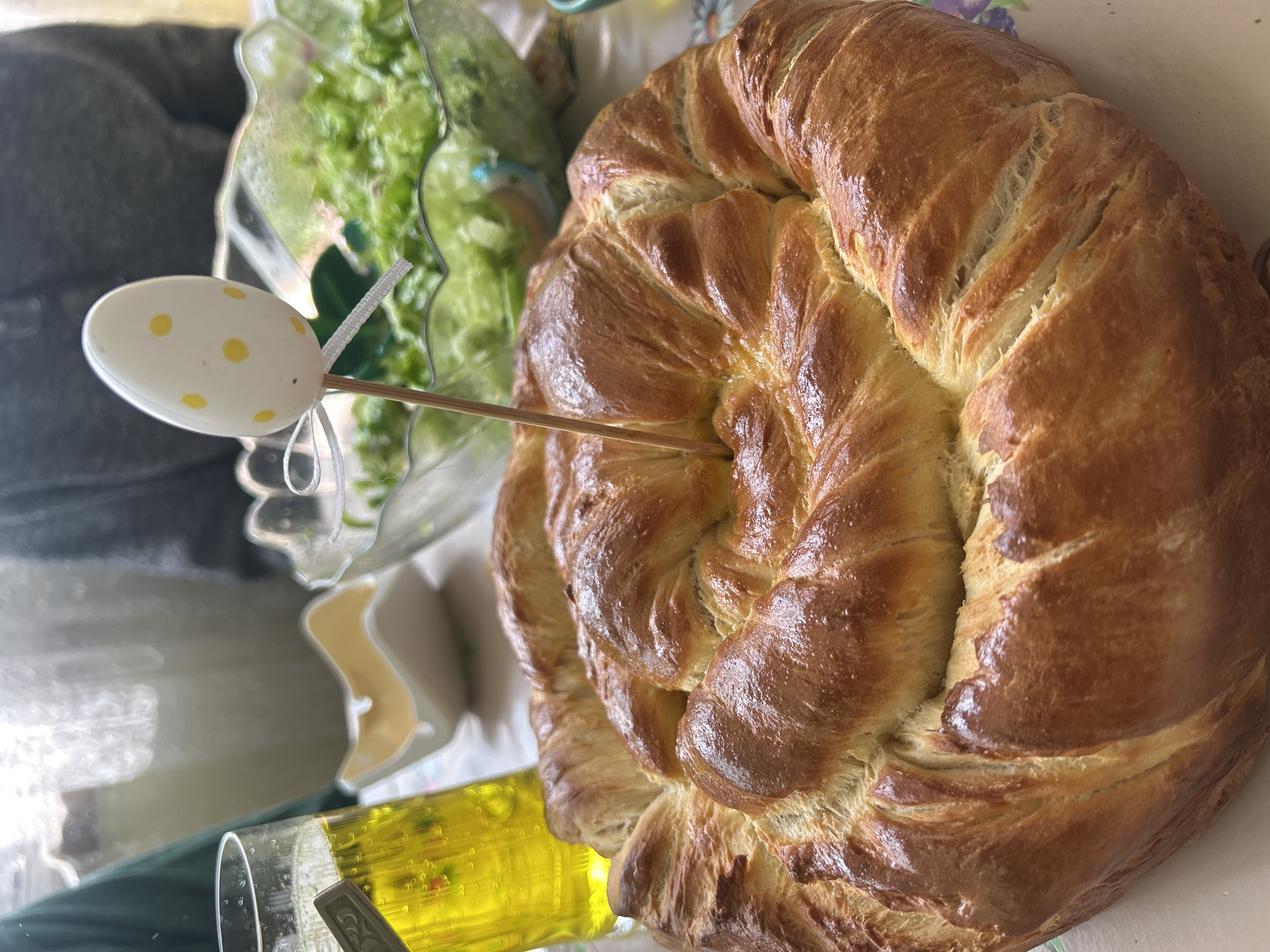 Here is a detailed description of the image:
The image presents a close-up shot of a braided bread loaf on a table. The loaf is golden-brown, suggesting it has been baked to perfection. A decorative white Easter egg with yellow dots on a wooden stick is inserted into the bread.
To the left of the bread, there's a glass bowl filled with green salad, adding a touch of freshness to the scene. A glass with a yellow liquid is near the bottom left, suggesting possibly olive oil. The table surface has a floral pattern, which provides a soft, decorative backdrop to the food items. The location is Unknown, Unknown.