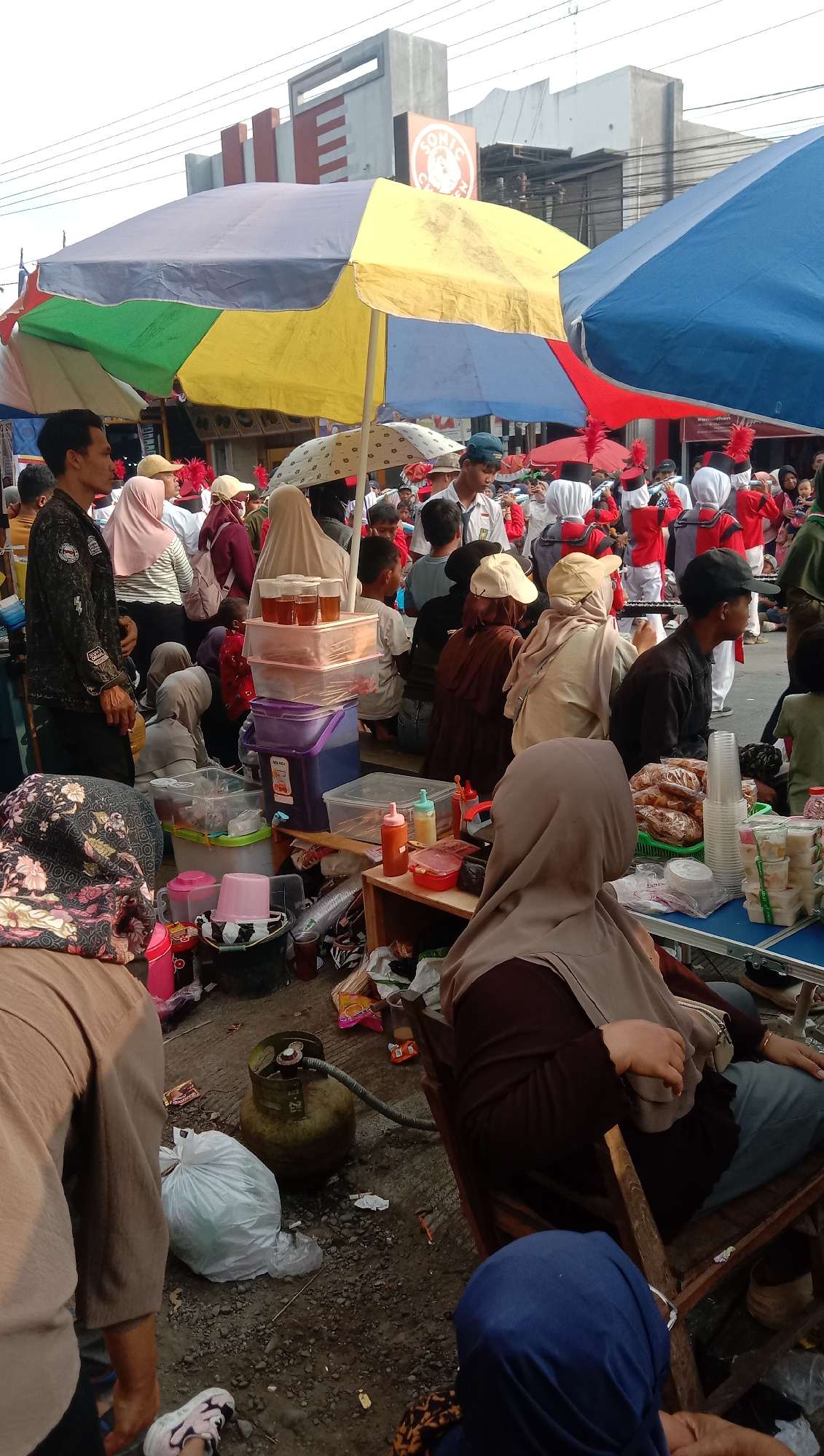 In Jatisawit, Indonesia, a crowded street market is depicted. A procession of people in red and white marching band uniforms with red feathered hats are moving through the scene, some playing instruments. They are surrounded by a crowd of onlookers. Various vendors are set up, with stalls displaying food and drinks. Several large, colorful umbrellas are providing shade over the market. In the foreground, a gas cylinder sits on the ground next to a plastic bag. The ground is a mix of dirt and gravel, with scattered trash. The overall impression is of a lively community event or festival.