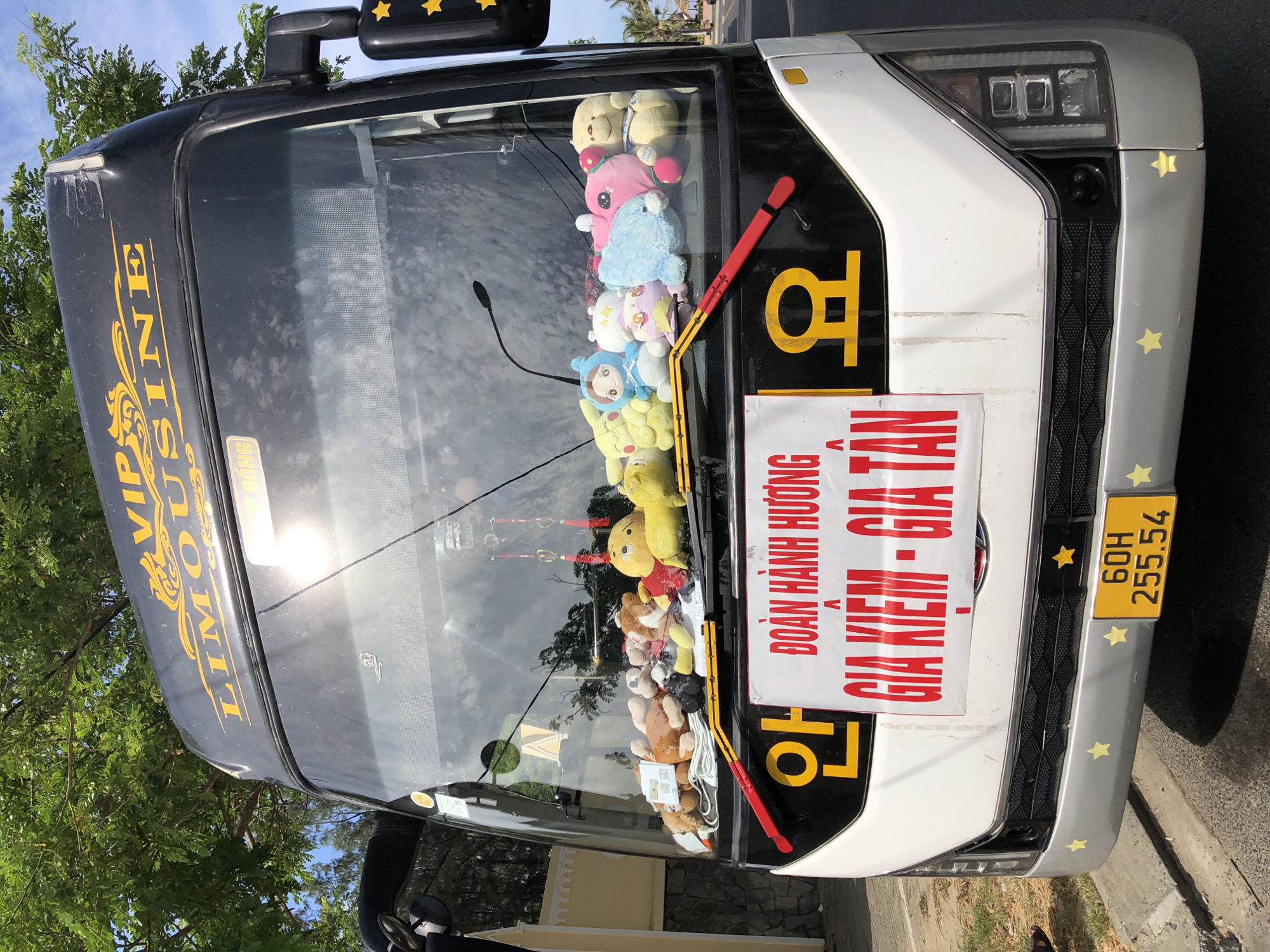 This is a medium shot of the front of a white and black VIP limousine bus parked on a paved surface. The bus has a license plate that reads "60H 255.54". The windshield is filled with numerous plush toys of various animals and characters. A red and white banner with Vietnamese text in red and yellow is affixed to the front of the bus, with large yellow characters to the left and right of the banner. The bus is parked outdoors, with green trees and a blue sky with clouds visible in the background. A glimpse of a road and palm trees suggests the location is likely in Đà Nẵng, Vietnam, possibly near a beach or coastal area.