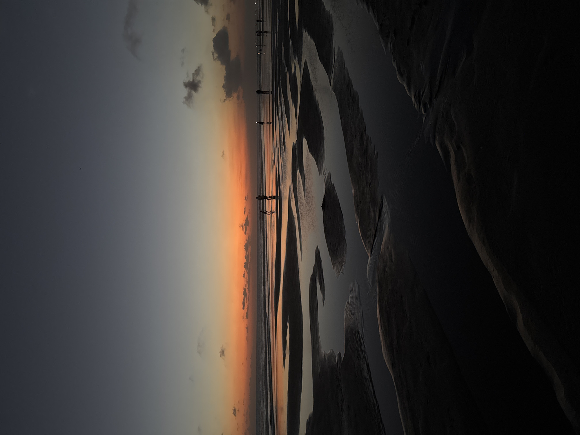This image depicts a beach scene at dusk, likely in Kuta, Indonesia, given the context. The low tide has exposed a textured sandy seabed with rippling patterns, reflecting the dusky sky. Several silhouetted figures of people are walking along the wet sand, their forms small against the vastness of the landscape. Some are walking individually, while others appear to be in small groups.
The sky transitions from a deep gray at the top to a warm, soft orange and yellow gradient near the horizon, indicating the sun has recently set. Scattered clouds are visible, some dark and dramatic, others lighter. The overall mood is serene and peaceful, characteristic of a sunset on a beach. The foreground is dominated by the dark, undulating shapes of the wet sand and shallow water, creating a stark contrast with the brighter, more distant horizon. There is no visible text in the image.