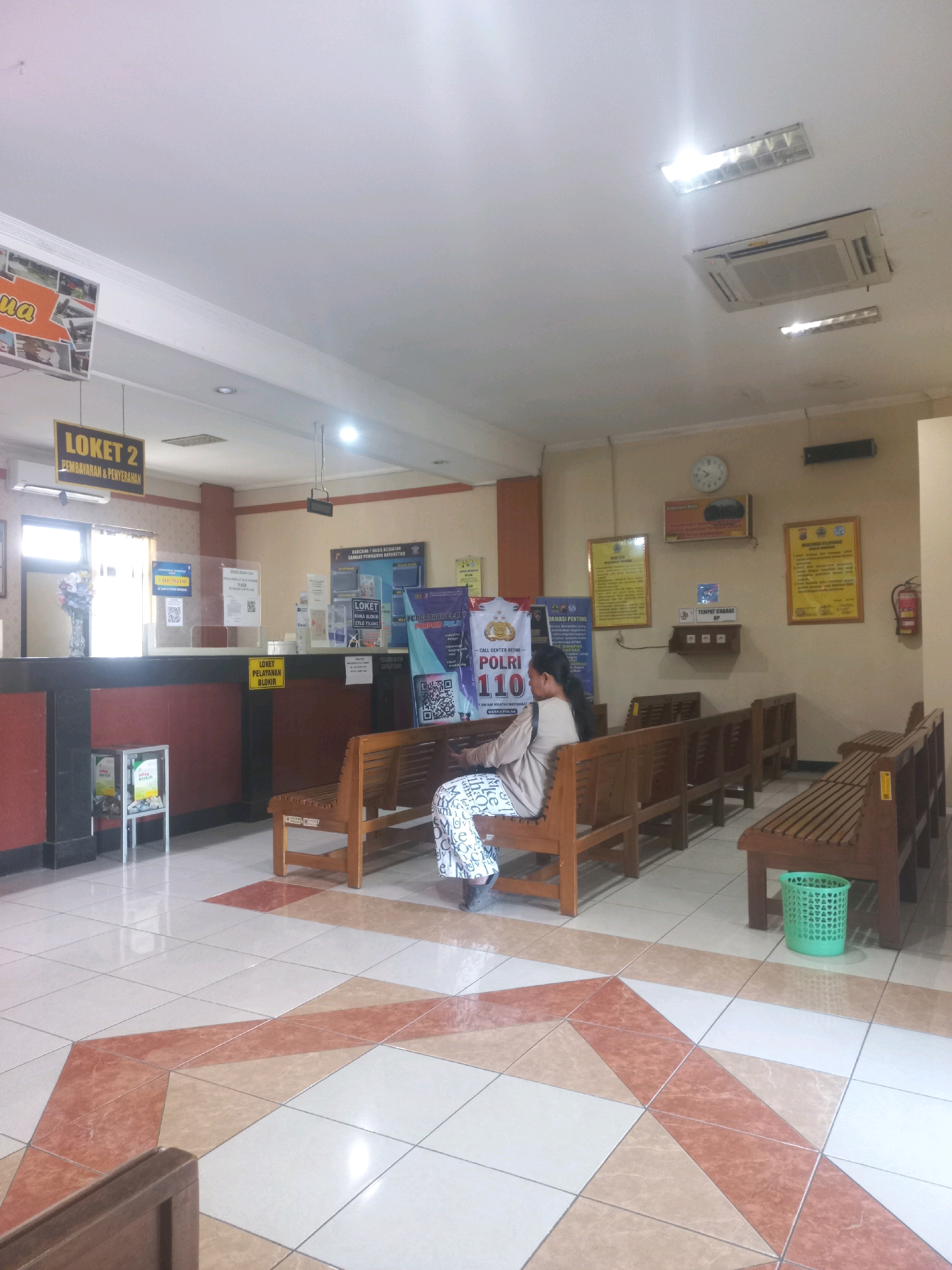 The image depicts the interior of what appears to be a public service office or waiting area in Baturetno, Indonesia. The scene is brightly lit by overhead fluorescent lights and an air conditioning unit is visible on the ceiling. The walls are painted a light beige.
In the foreground, a woman is seated on a wooden bench, facing away from the camera. She is wearing a light-colored patterned garment. Several other empty wooden benches are arranged in rows, suggesting a waiting area. The floor is tiled with a geometric pattern of light beige and reddish-brown tiles. A green plastic trash bin is positioned near the benches.
In the background, there is a counter area with various signs and informational posters displayed on the wall. One prominent sign reads "LOKET 2 PEMBAYARAN & PENYERAHAN" (Counter 2 Payment & Submission). There are also posters related to police services, including one with the number "110" and "POLRI" (Indonesian National Police). A glass partition is present at the counter, and various items, possibly for service or information, are visible behind it. A fire extinguisher is mounted on the wall to the right. A clock is visible on the wall above a poster.