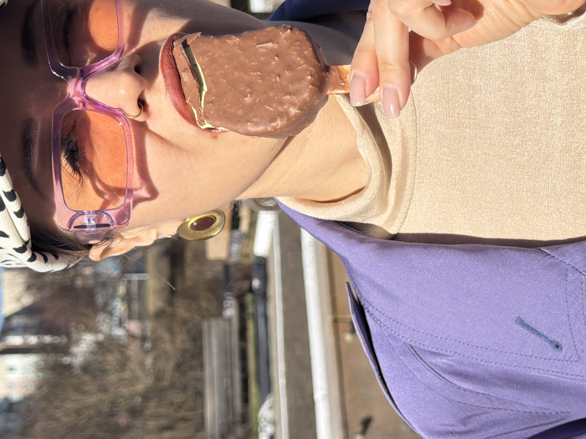 A close-up shot, rotated counter-clockwise, captures a person eating an ice cream bar outdoors in Krasnoyarsk, Russia. The individual, depicted from the chest up, is seen holding a chocolate-covered ice cream bar with a visible green filling, possibly mint, up to their mouth. Their lips are adorned with a dark reddish-brown lipstick.
The person is wearing large, transparent pink-framed sunglasses with orange-tinted lenses, obscuring their eyes. A small gold nose ring is visible on their right nostril, and a larger gold, circular earring hangs from their right ear. They are dressed in a light beige top, layered under a purple jacket or shirt, and a black and white patterned head covering or bandana is visible on the left side of their head. The ice cream bar is held by a hand with natural, unpolished nails.
The background is softly blurred, suggesting an outdoor setting with hints of trees or foliage and distant structures. The bright, direct sunlight indicates a clear, sunny daytime environment. No specific emotions are discernible due to the sunglasses covering the eyes, and there is no visible text in the image.