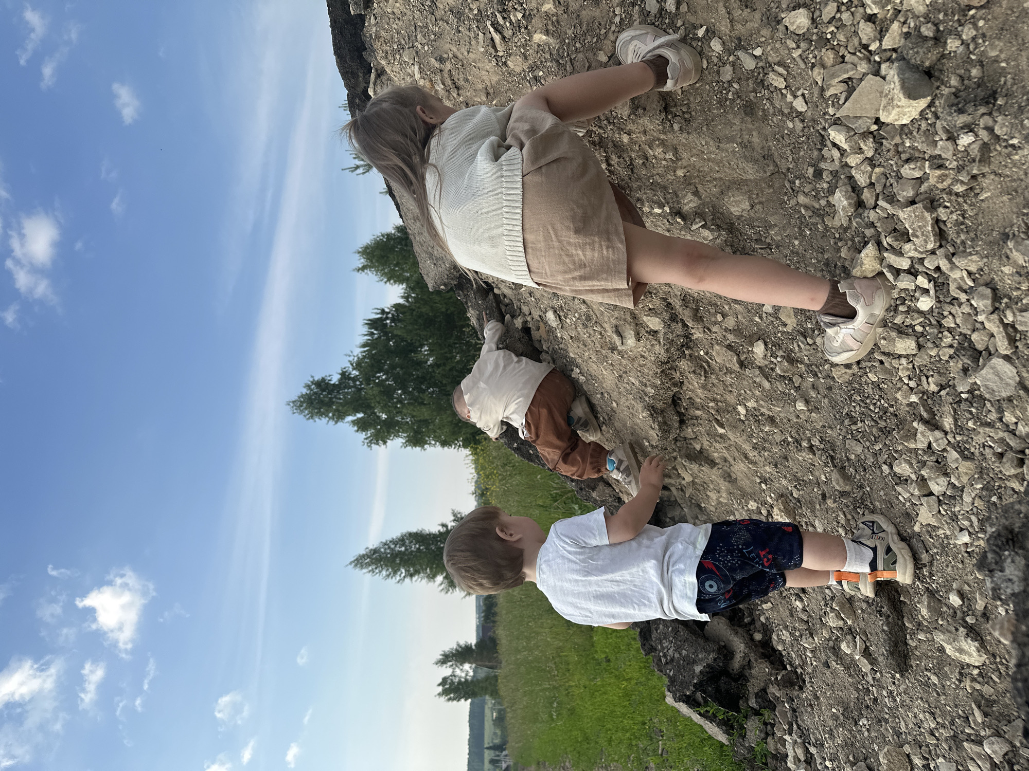 Three children are on a dirt hill under a partly cloudy blue sky. In the foreground, a young boy wearing a white t-shirt and patterned shorts stands looking to his right. Behind him and to his right, a younger child in brown pants and a white shirt is crawling up the hill. To the right of the image, a girl wearing a cream-colored sweater and a beige skirt is climbing the hill, facing away from the camera. The hill is composed of dirt and rocks, with some debris that appears to be asphalt. In the background, there is a green field, trees, and a hint of distant buildings. The setting appears to be a rural or semi-rural area outside of Эстачи, Russia.
