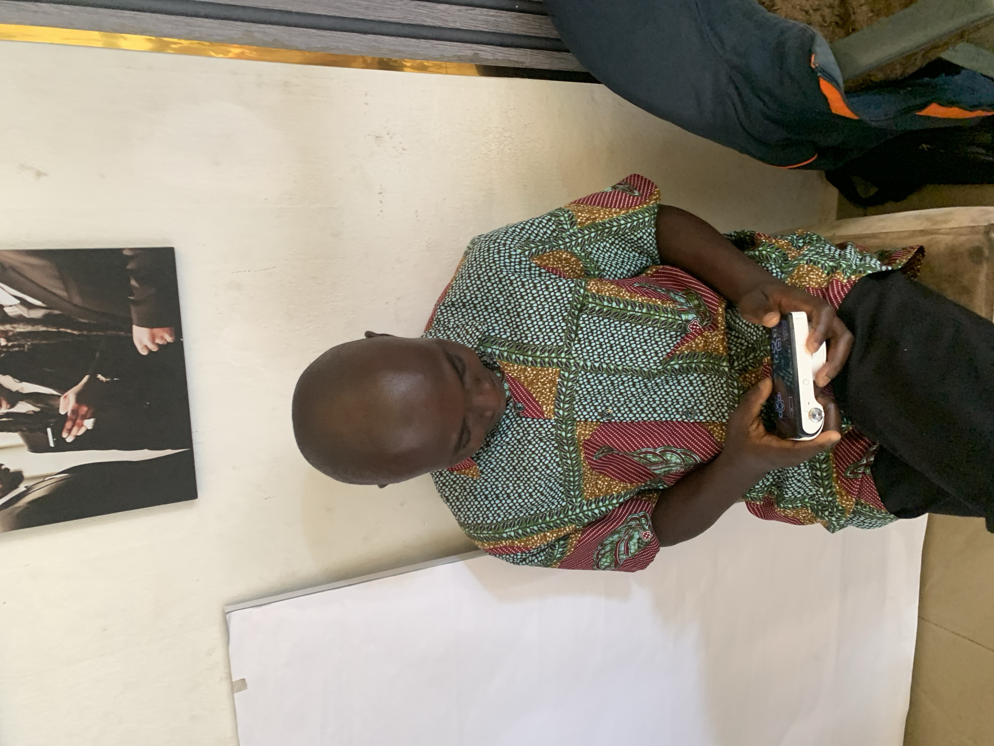 A bald Black man wearing a colorful patterned short-sleeved shirt and black pants is seated and looking down at a small white electronic device he is holding with both hands. The man is positioned in what appears to be an indoor setting with a light-colored textured wall behind him. To his left is a large white board or easel. To his right, a dark-colored leg with an orange stripe on the pant leg is visible, suggesting another person is present but out of frame. The man's shirt features a repeating pattern of geometric shapes and lines in shades of blue, green, red, and yellow. The device he is holding is rectangular with a screen and some buttons. Above the man, a framed picture is hanging on the wall, depicting two men in suits.