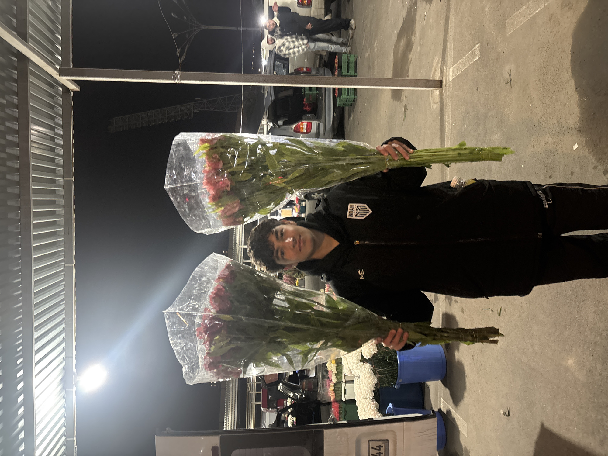 A person holds two large bunches of red flowers in plastic wrap in an outdoor setting at night. The person is wearing a black jacket with a logo on the chest. In the background, a vehicle is partially visible with other people standing near it. A construction crane is visible in the distance. Other flower displays and containers are seen in the scene. A possible license plate can be seen on the vehicle. While a positive ID is not possible, the setting, appearance of the possible license plate, and background details suggest the location might be a flower market in Yerevan, Armenia.