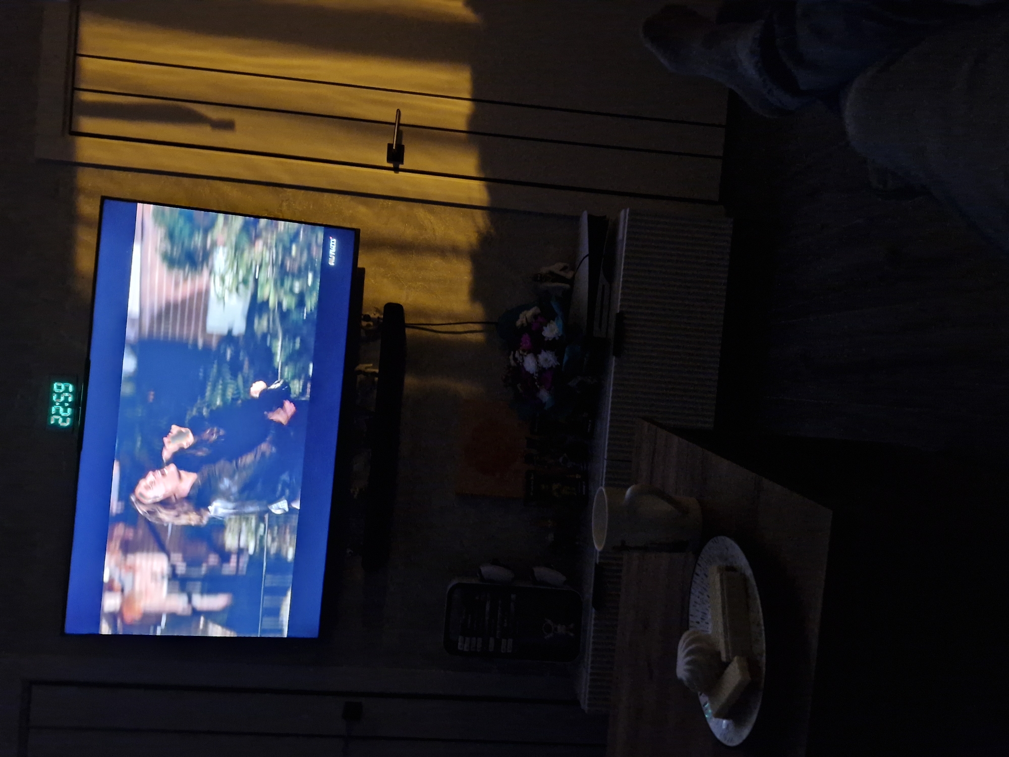 The image depicts a dimly lit living room interior, with a large television screen displaying a scene of two individuals, one embracing the other. A digital clock on top of the television displays the time as 22:59. Below the television, on a shelf, are various electronic devices, a mug, and a bouquet of flowers. In the foreground, a plate with two pieces of cake or pastry and a marshmallow sits on a wooden surface, possibly a coffee table. The overall lighting suggests it is nighttime, with a strong yellow light casting shadows on the right side of the image, possibly from an external source or a lamp. The scene is indoors, likely a residential setting, and there is no indication of its specific location within the city of Kemerovo, Russia, or any other specific geographic area. The image captures a moment of relaxation or entertainment, with the focus on the television screen and the nearby snacks.