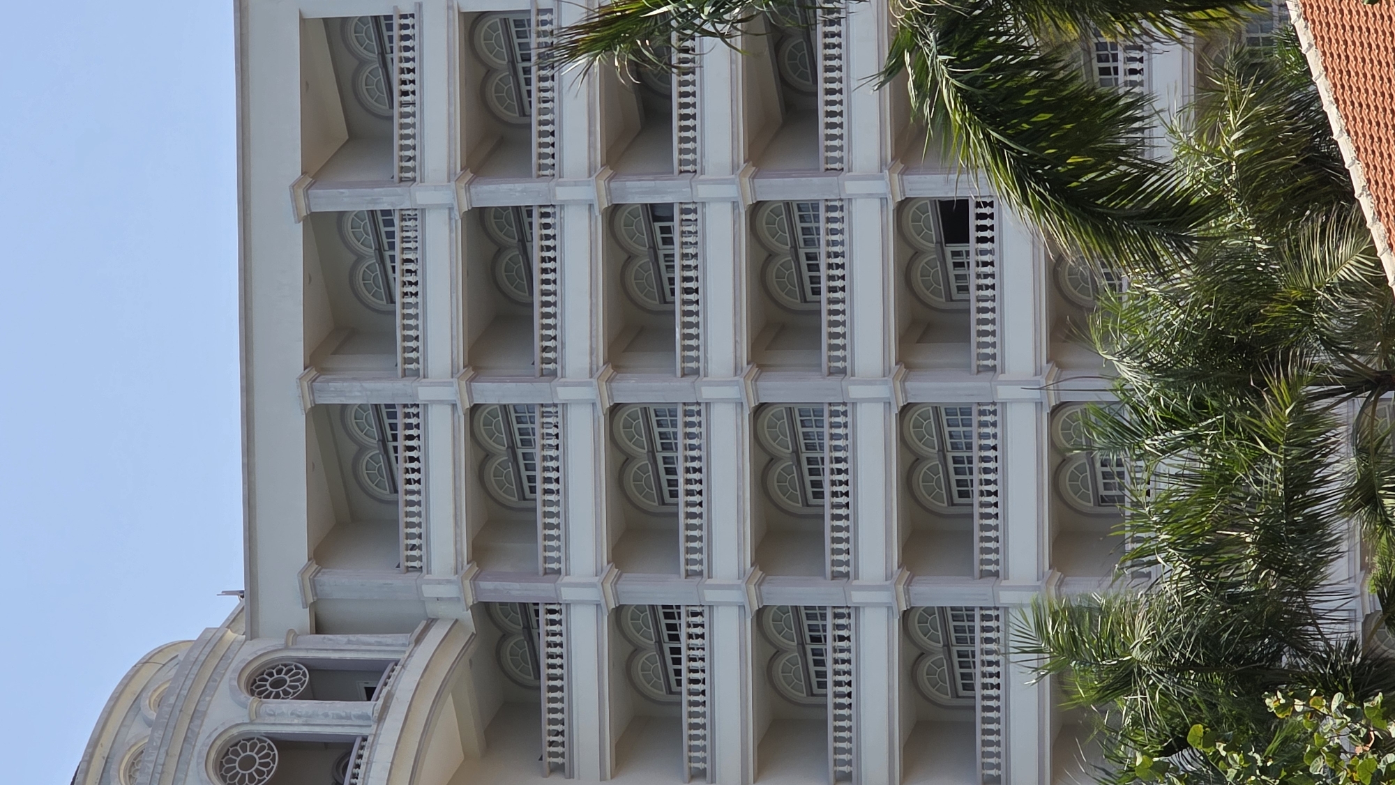 A tall, light-colored building with multiple rows of balconies is partially obscured by palm trees. The balconies feature arched windows or openings and detailed balustrades. The structure exhibits classical architectural elements. The location is possibly Nha Trang, Vietnam.