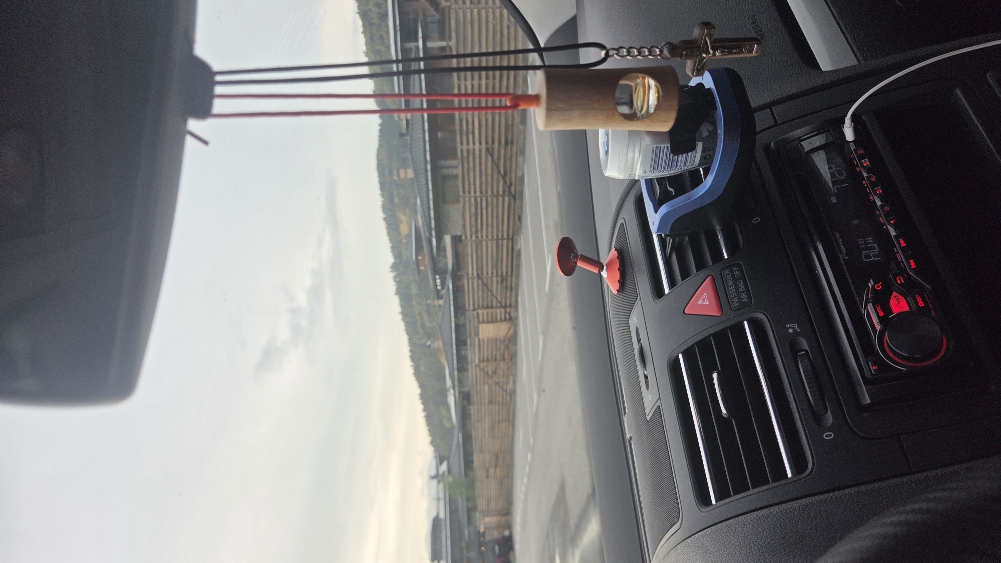 This is a close-up, interior view of a car's dashboard, facing forward through the windshield. The focus is on the dashboard's air vents, car stereo, and a blue cup holder attachment. A red emergency hazard light button is visible above the stereo. Hanging from the rearview mirror are two strings, one red and one black, with a wooden air freshener and a crucifix attached to the black string. A white USB cable is plugged into the car stereo. Outside the car, a parking lot is visible, bordered by a wooden fence and buildings. The sky is overcast.