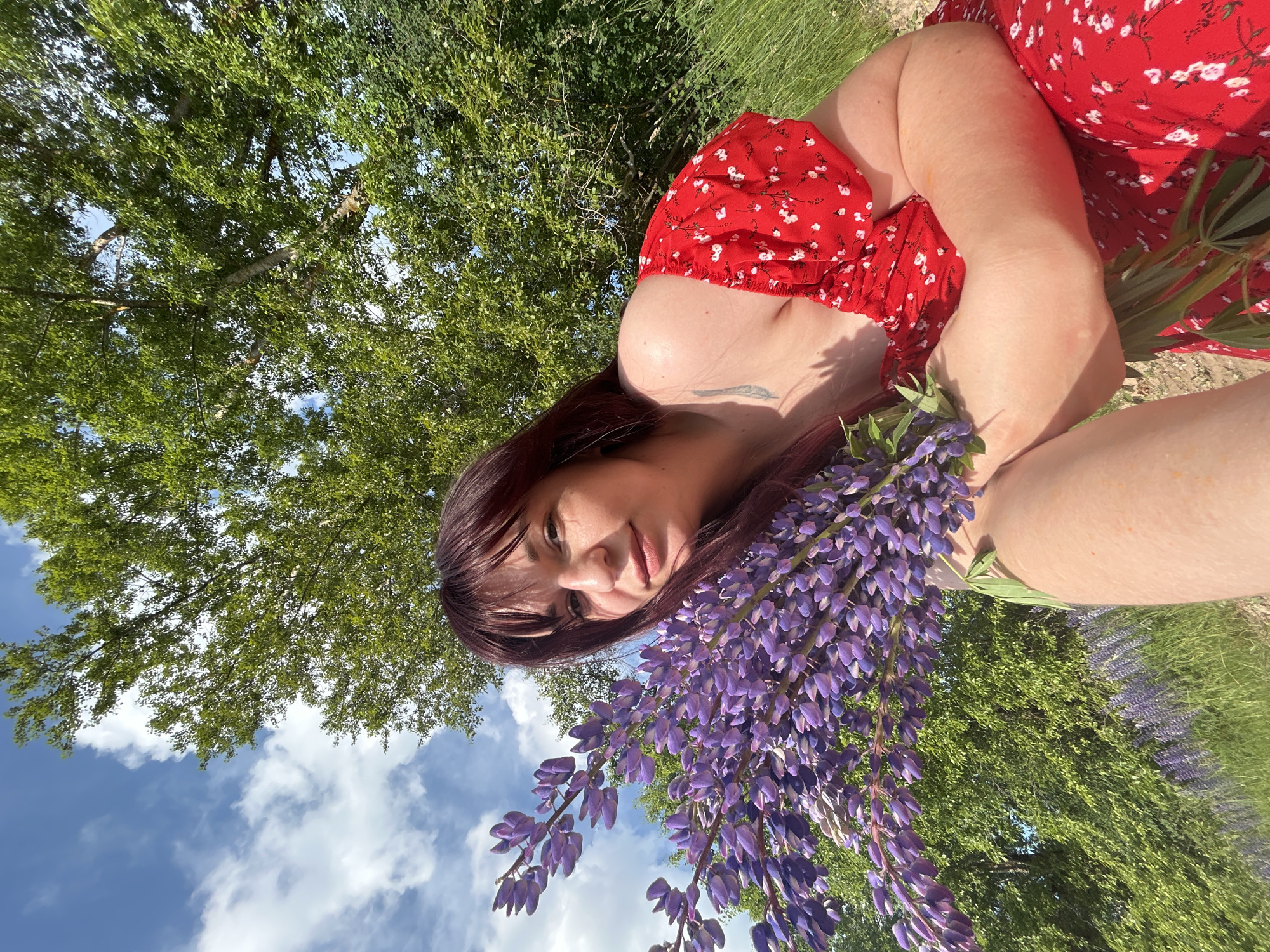 A woman with long, dark red hair and bangs is holding a large bouquet of purple lupine flowers. She is wearing a red dress with a white floral pattern. The woman is outdoors in a grassy field with trees and a blue sky with white clouds in the background. In the distance, more purple lupine flowers are visible. The location is stated to be in the city of Goryachevo, Russia.