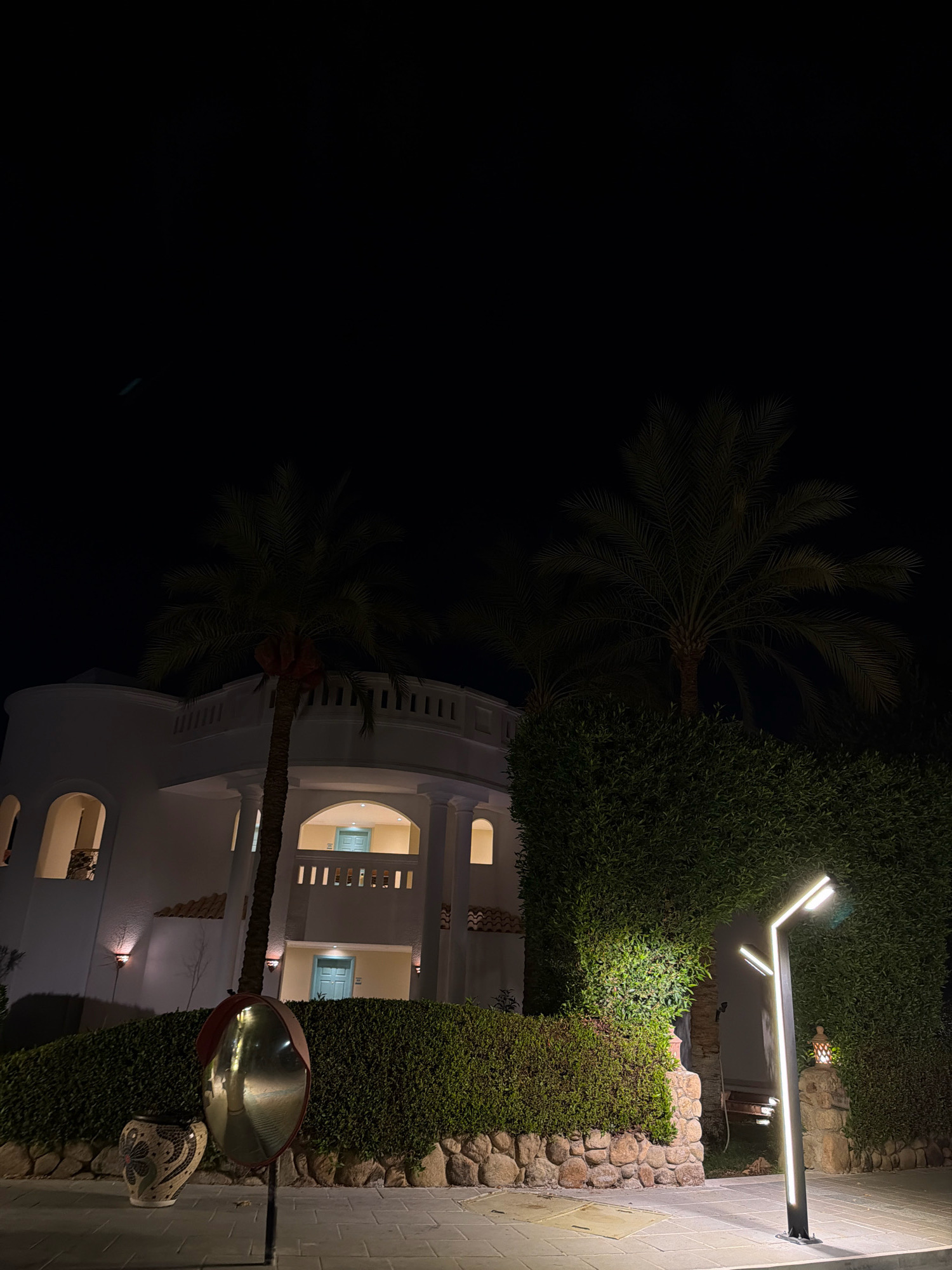 At night in Sharm El Sheikh, Egypt, a well-lit outdoor scene features a modern white building with arched windows and balconies, flanked by lush green hedges. Two palm trees are visible, one in the foreground and another in the background. In the foreground, a decorative vase and a convex mirror mounted on a pole are present. A contemporary street lamp with multiple illuminated sections stands to the right, casting light on the paved ground. The sky is dark, indicating nighttime.