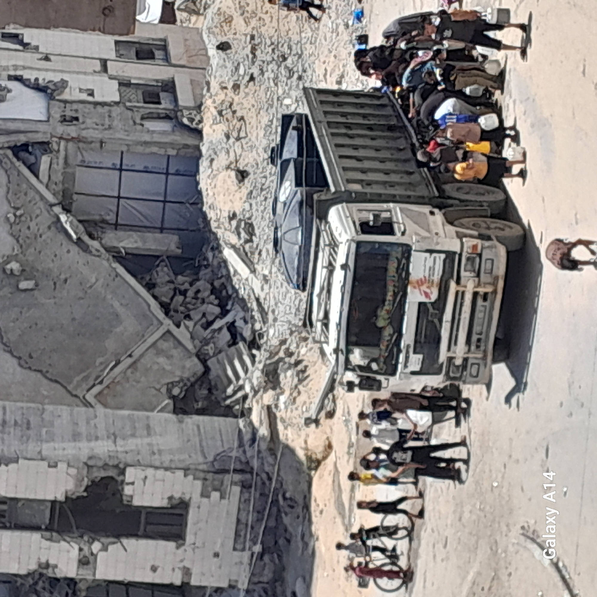 In the city of Khan Yunis, Palestinian Territories, a heavy-duty truck is parked on a dusty, rubble-strewn road in front of a damaged building. The building, with its concrete structure exposed and windows boarded up or broken, appears to have suffered significant destruction.
A group of people, including men, women, and children, are gathered around the back of the truck. Some are carrying white plastic water containers, suggesting they are collecting or distributing water. The truck itself has two large black water tanks on its roof. The front of the truck displays logos, one appearing to be from "Medecins Sans Frontieres."
To the left of the truck, several children are seen near bicycles. The ground is covered in sand and debris, indicative of a damaged urban environment. The scene conveys a sense of urgency and resource distribution, possibly in response to a crisis.
