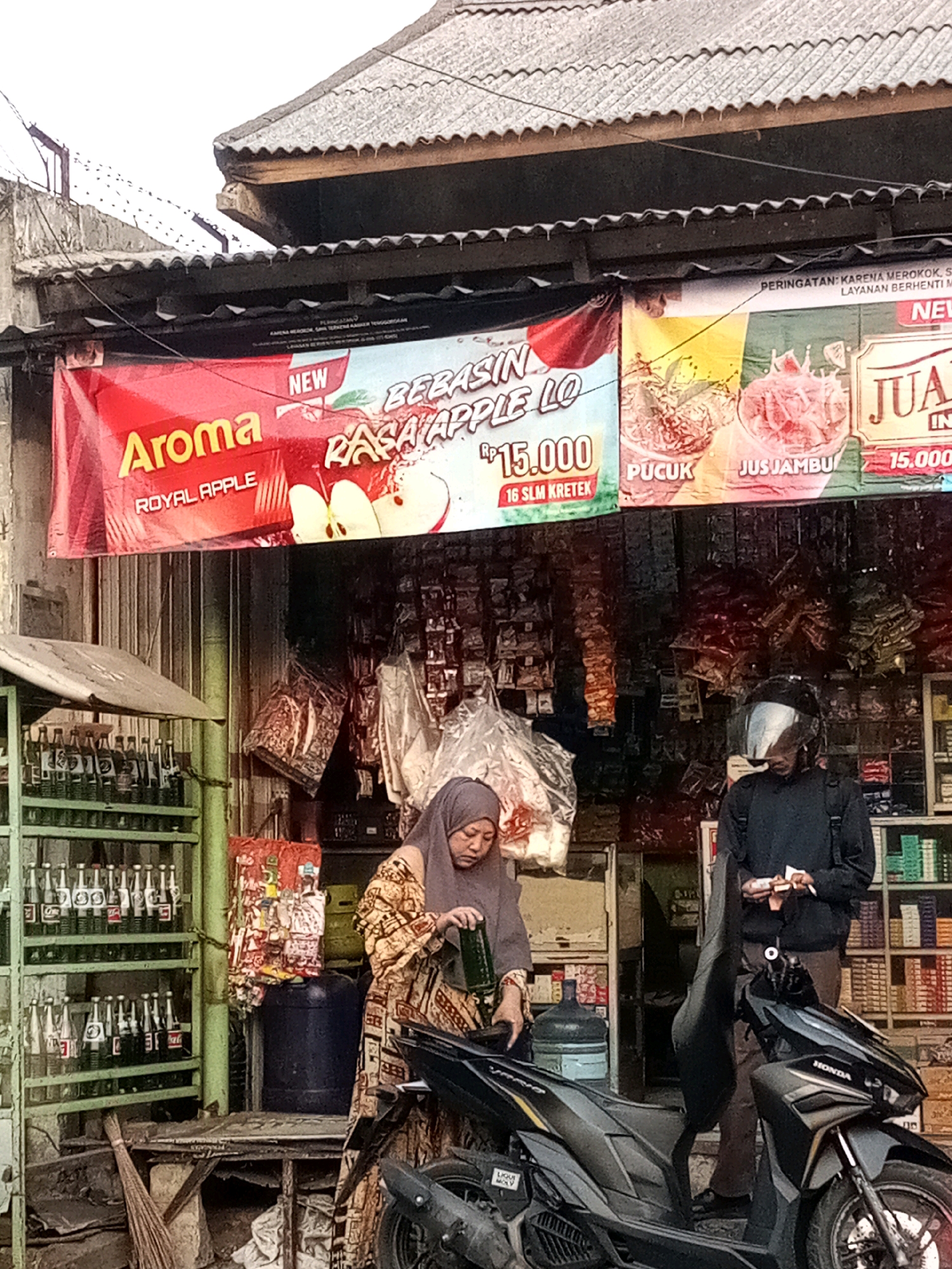 The image depicts a street-level view of a small convenience store in Bekasi, Indonesia, likely in an urban or semi-urban setting, indicated by the presence of a tiled roof, corrugated metal sheeting, and an overhead wire. The store displays various goods, including bottled beverages on a green metal rack, packaged cigarette cartons on shelves behind the counter, and what appear to be loose packets of goods hanging from the ceiling. Two individuals are present: a woman in the foreground, wearing a hijab and patterned traditional clothing, is attending to something near a large blue plastic barrel. She is partially obscured by a parked black scooter, identified by the "HONDA" logo. To her right, a man wearing a helmet, dark jacket, and pants stands behind the counter, holding a small item, possibly a receipt or money. The store has a prominent red banner advertising "Aroma Royal Apple" with an apple graphic and "BEBAS Rasa Apple Lo Rp.15.000" and another banner for "JUS JAMBU 15.000". The overall scene suggests a typical local shop operating in Indonesia, with a focus on readily available consumer goods. The time of day appears to be late afternoon or early evening, given the soft, diffused lighting.