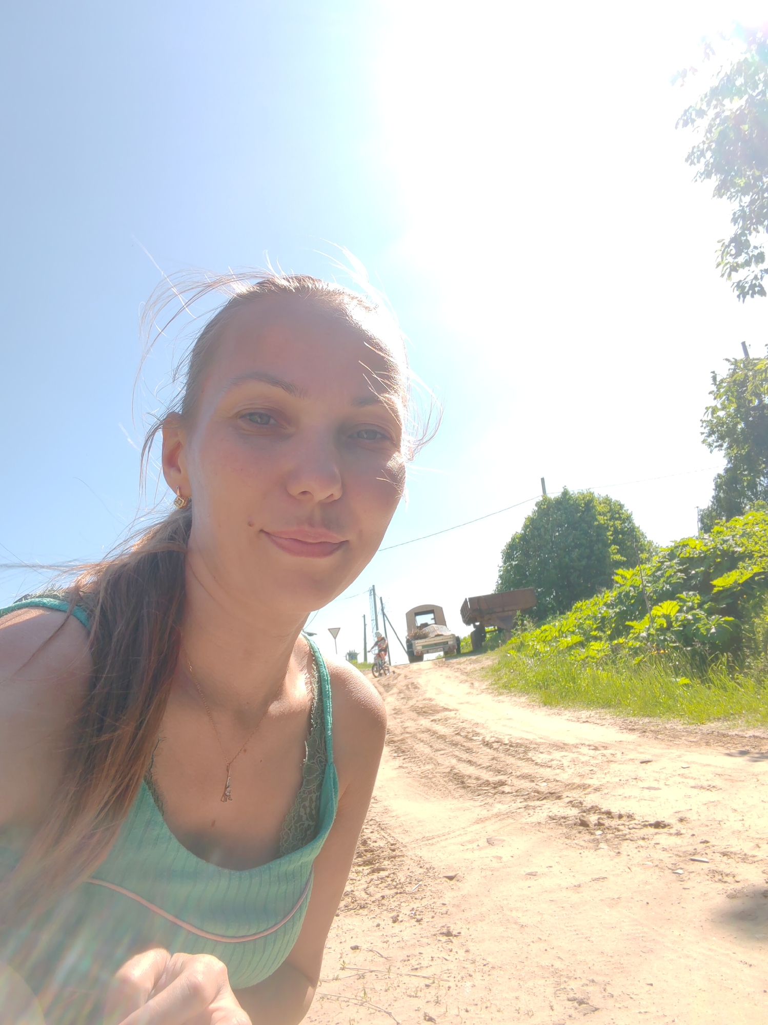 A young woman with long, light brown hair pulled back in a ponytail takes a selfie outdoors on a sunny day. She is wearing a teal tank top with lace trim and a gold necklace with a pendant. Her left arm is partially visible in the foreground, with her fingers extended. Behind her, a dirt road curves into the distance. A small, older model truck and a trailer are parked on the side of the road, with a person on a bicycle visible further down the road. Lush green foliage and trees line the right side of the road, with a bright sky overhead. The scene appears to be in a rural or semi-rural area, possibly in or near the village of Chuchlam, Russia, given the style of the vehicles and vegetation.