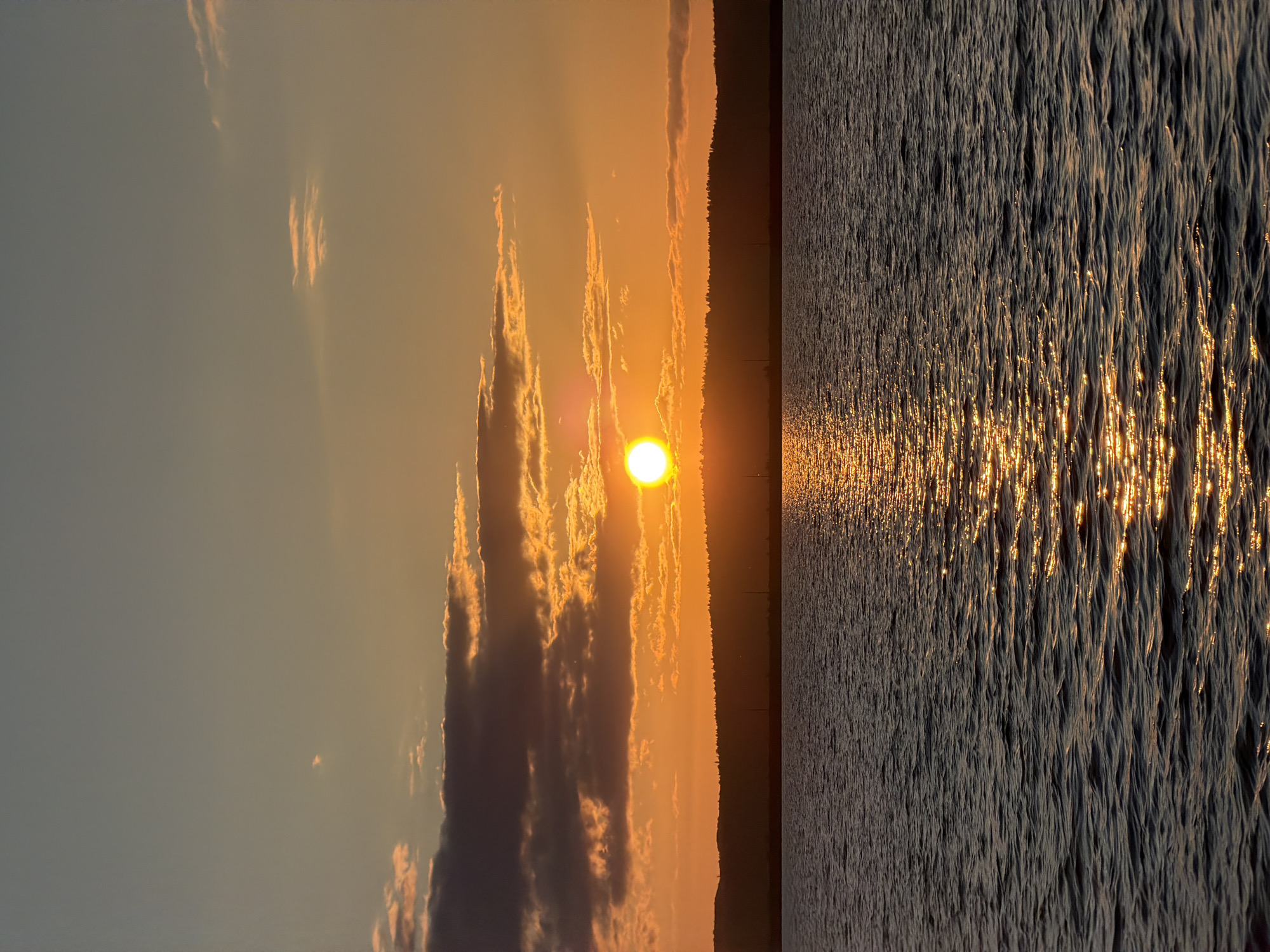 The image depicts a sunset over a body of water, likely the Kama River, in Chaikovsky, Russia. The sun, a bright orb, is partially obscured by clouds, casting a warm orange glow across the sky. Silhouetted against the vibrant sky is a tree-lined hill, its dark form creating a stark contrast to the bright hues. The water in the foreground reflects the sunset, creating a shimmering path of light that stretches towards the viewer. The water is slightly rippled, suggesting a gentle breeze. The overall scene is one of natural beauty and tranquility.