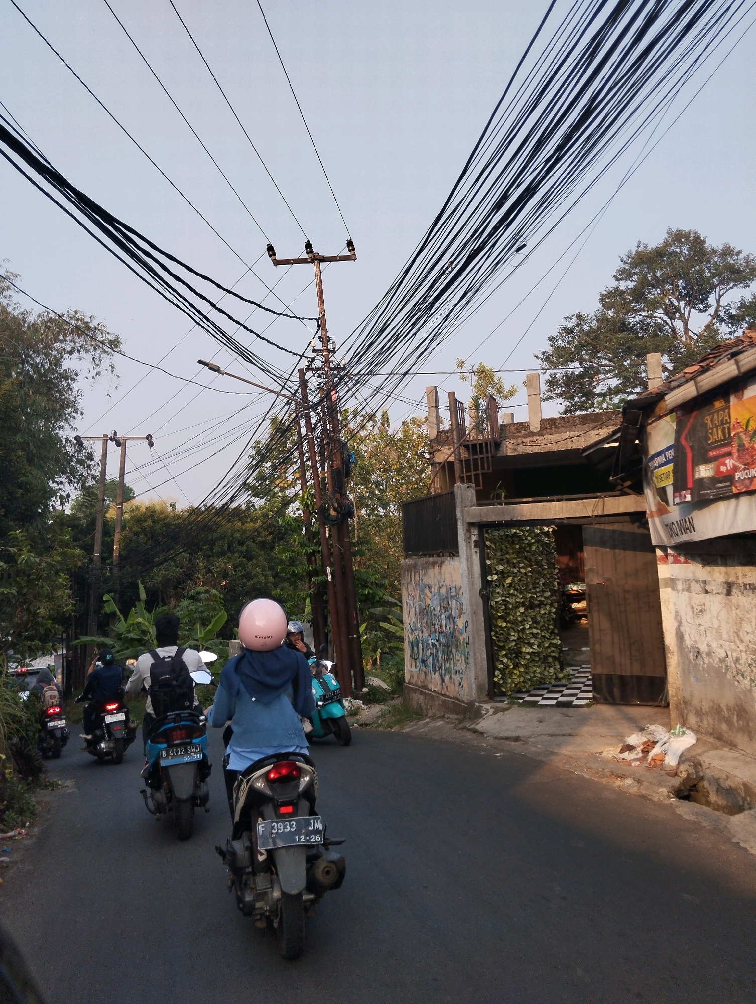 This image captures a street scene in Sukaraja, Indonesia, characterized by a dense overhead network of electrical wires crisscrossing the sky. In the foreground, a group of people are riding motorcycles on a paved road. The motorcycle closest to the viewer has a license plate reading "F 3933 JM 12-26" and the rider is wearing a blue hoodie and a pink helmet. Behind them, another motorcycle rider with a backpack is visible. Further down the road, more motorcycles and riders are in motion. The roadside is lined with trees and vegetation, with a building visible on the right side of the frame. This building features graffiti on its lower section and a dark entrance covered partially by artificial greenery. A sign advertising "KAPA SAKT" is affixed to the building's exterior. The sky is overcast, and the overall atmosphere suggests an urban or semi-urban environment with visible infrastructure and daily life activities.