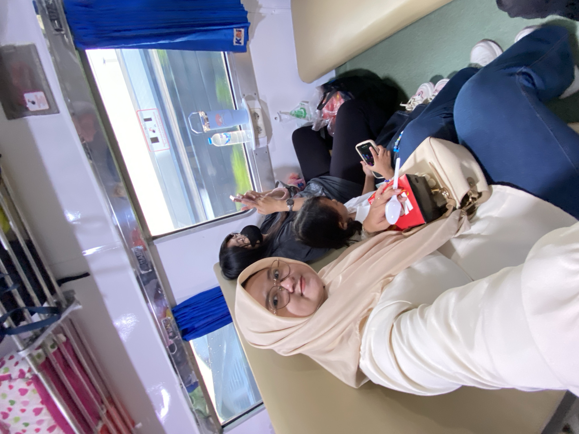 The image shows several people sitting in a train car. At least one window is visible with a blue curtain. A woman wearing a hijab and glasses is in the foreground. Other passengers are visible, some using mobile phones. Water bottles are placed on the wall beside the window.
The KAI logo is visible above the curtain, a common indicator of Indonesian trains. The origin place, Tenggulunan, is not discernible from the provided visual data.