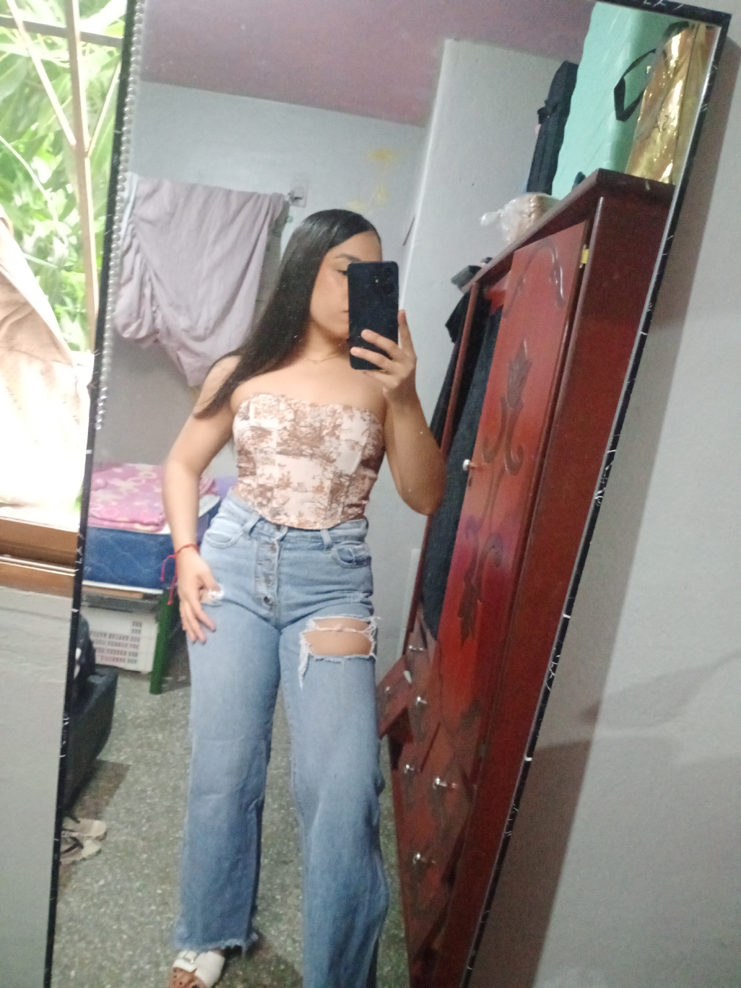 A young woman is taking a selfie in a mirror, presumably in an indoor setting in Valle de La Pascua, Venezuela. She is wearing a strapless patterned top and ripped jeans. Her face is partially obscured by her phone. Behind her, there is a bed with multiple blankets and pillows, a wardrobe, and a window showing foliage outside.