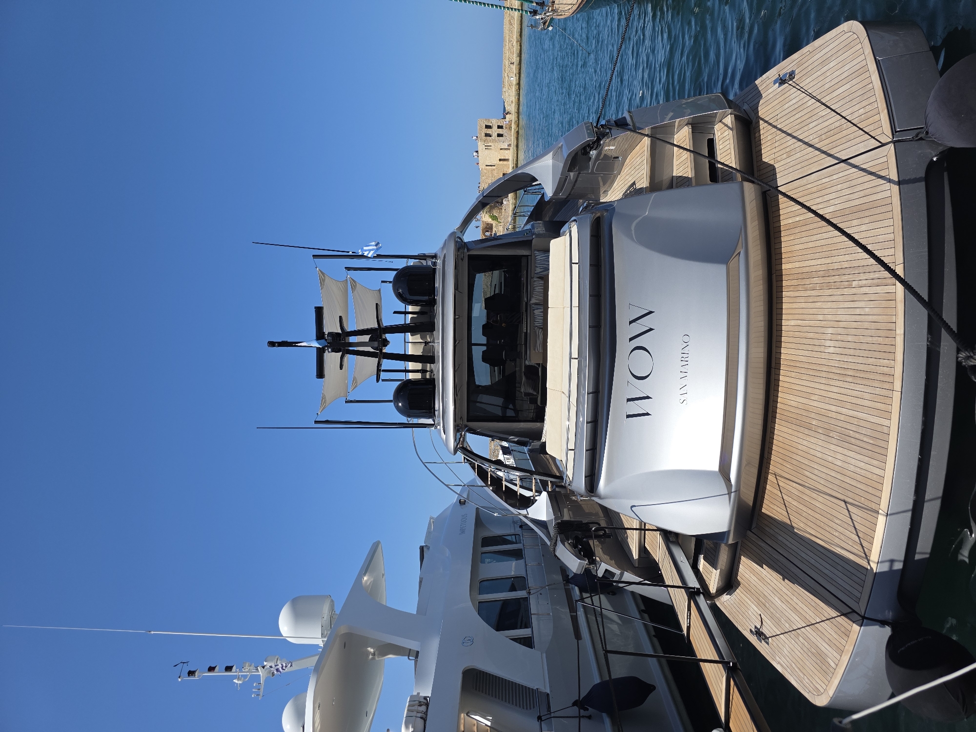 This image shows two yachts moored in the Venetian Harbor of Chania, Greece. The foreground features a sleek, silver yacht with "WOW" and "SAN MARINO" printed on its stern. The yacht has a wooden deck and a dark hull. Behind it, a larger white yacht, identified by the text "IMPETUOUS" on its side, is also docked. The harbor water is a clear blue, reflecting the bright, cloudless sky. In the background, the historic Venetian fortress and stone walls of the harbor are visible, with a few people standing on top of them. The scene is bathed in sunlight, indicating a clear day.