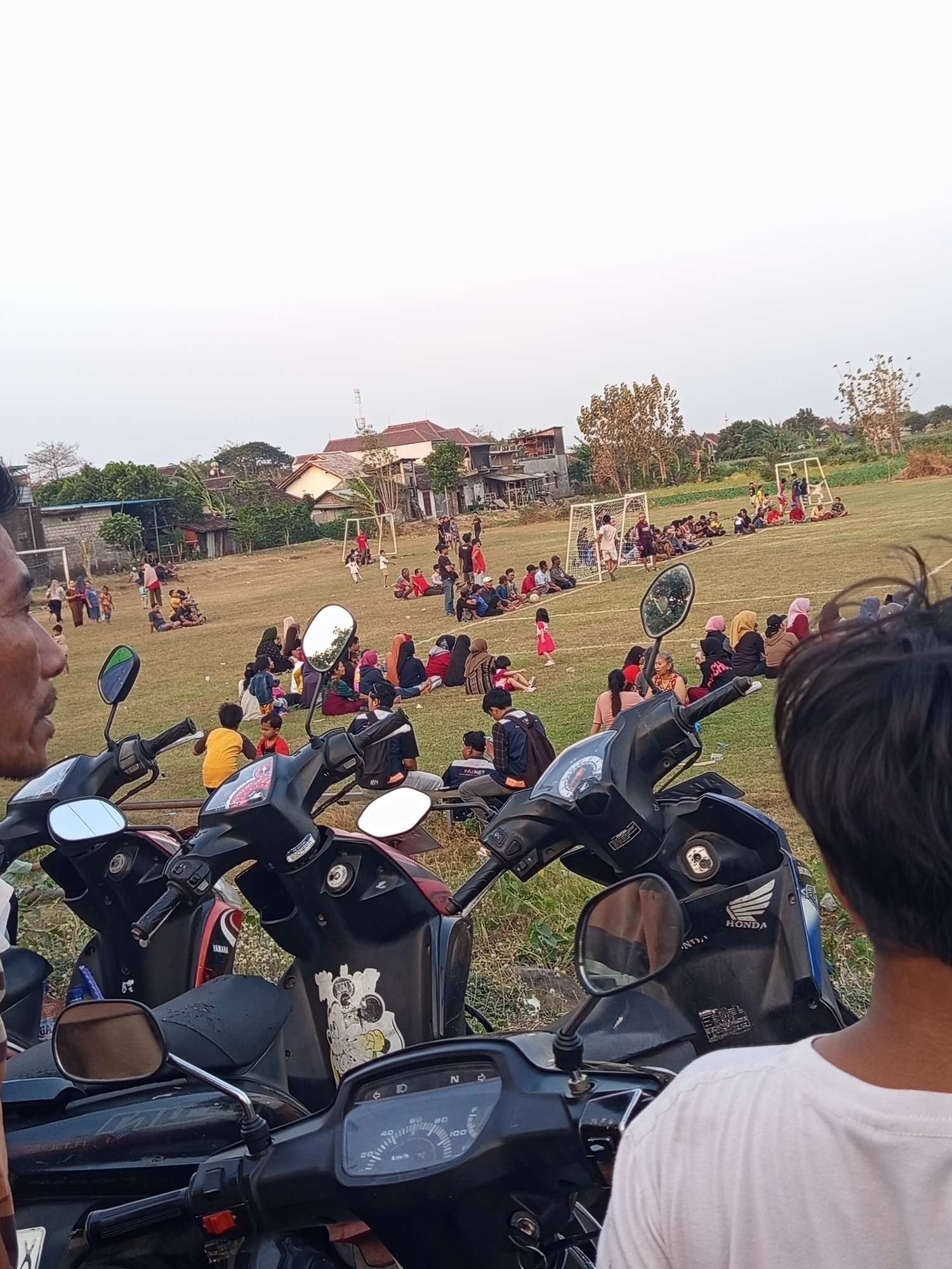 The image depicts an outdoor scene in Mrican, Indonesia, likely a village or a rural outskirts, as suggested by the presence of fields, scattered houses, and a community gathering. In the foreground, several motorcycles are parked, their handlebars and mirrors visible. The focus then shifts to a grassy field where a group of people, including men, women, and children, are gathered. Some are seated on the grass, while others appear to be watching an activity. A soccer goal is visible in the background, suggesting a game or practice might be taking place. The people are dressed in casual attire, with some women wearing headscarves. The overall atmosphere appears to be relaxed, with a sense of community engagement. The environment is characterized by a muted color palette, with the green of the grass, the earthy tones of the houses, and the pale sky.