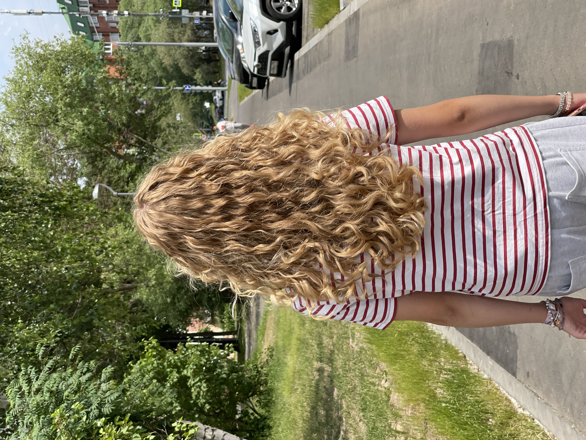 The image shows a young person with long, curly blonde hair from behind, walking on a paved sidewalk next to a grassy area. They are wearing a red and white striped t-shirt and grey sweatpants, with several bracelets on their left wrist. In the background, there are trees, buildings, parked cars, and street signs. The scene appears to be a residential or commercial street in a city, likely Moscow, Russia, given the presence of distinctive architecture and street signage, such as the blue and white traffic signs. The lighting suggests it is daytime with clear skies.