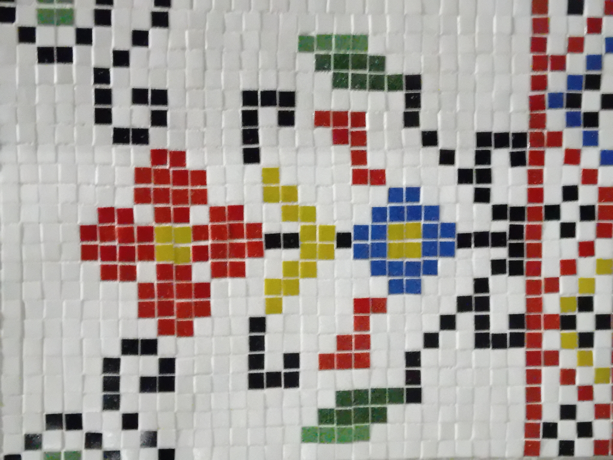 The image displays a close-up view of a vibrant mosaic artwork composed of small, square tiles. The primary subject is a decorative pattern featuring stylized floral and organic motifs against a white background.
No activity or event is taking place, as the image captures a static architectural or artistic surface.
Notable details include the colorful design, which incorporates red flowers with yellow centers, blue flowers also with yellow centers, and green tiles forming leaves or stems. Black tiles are used extensively for outlining shapes, creating additional abstract stem-like elements, and connecting the various floral components. The arrangement of the square tiles gives the mosaic a pixelated aesthetic, akin to cross-stitch patterns or early digital graphics. The tiles appear to be glossy, possibly ceramic or glass, exhibiting a subtle sheen. The individual tiles and their grout lines are clearly visible, adding texture to the surface. The image is captured at a slight angle, allowing for a perspective on the tiled surface. There are no visible location cues beyond it being a tiled surface, and no text is present in the image.