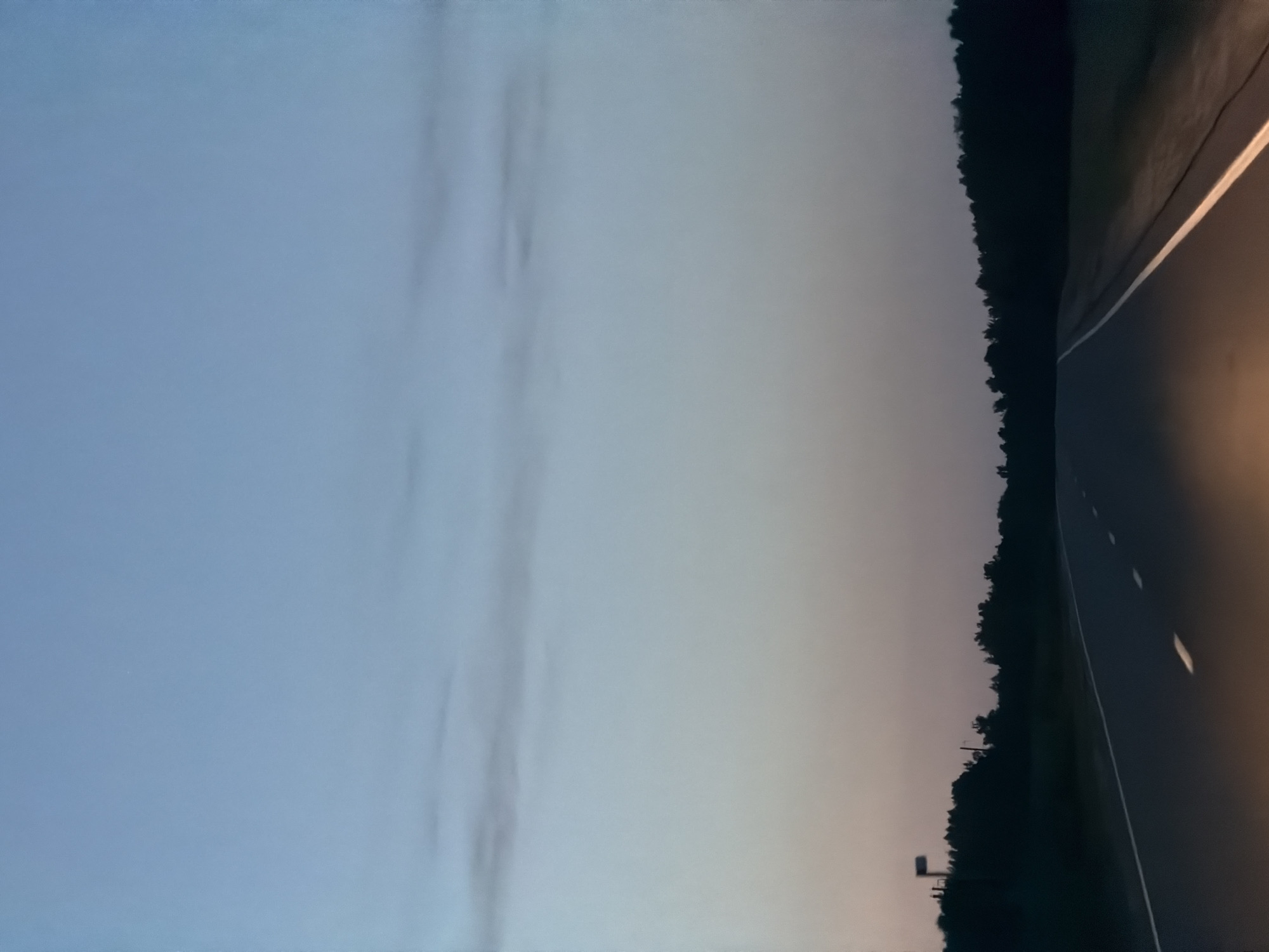 The image depicts a straight, two-lane asphalt road stretching into the distance under a twilight sky. The sky transitions from a pale blue at the top to a gradient of pink and gray near the horizon. A dark line of trees borders the right side of the road, with a lighter silhouette of trees and a single signpost visible on the left. The road has white dashed lines marking the lanes, and the headlights of a vehicle are visible in the foreground, illuminating a portion of the asphalt. The scene appears to be a rural or semi-rural area. The location is not specified within the image itself; therefore, a specific location within Laishevo, Russia, cannot be identified.
