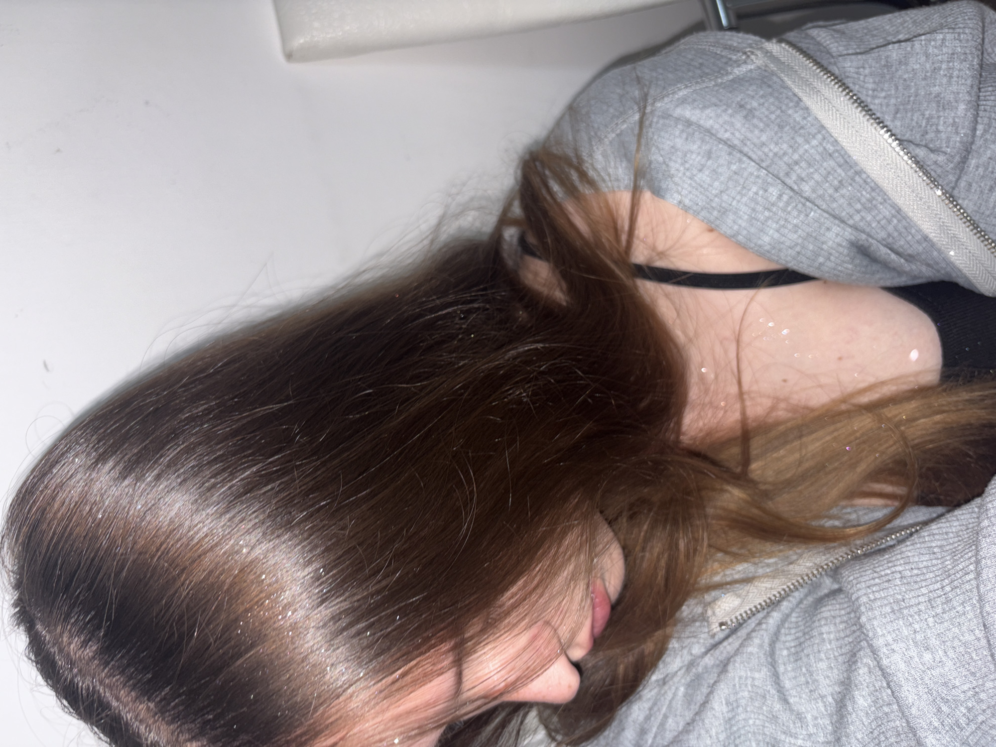The image shows a young woman from the shoulders up, looking down and to the side. Her hair is long and brown, with some glitter in it. She is wearing a grey zip-up hoodie over a black bra. The background is a plain white wall. The lighting is soft, with a slight glow from an unseen source. The image does not contain any information that would indicate it was taken in Chaikovsky, Russia, or any other specific location.