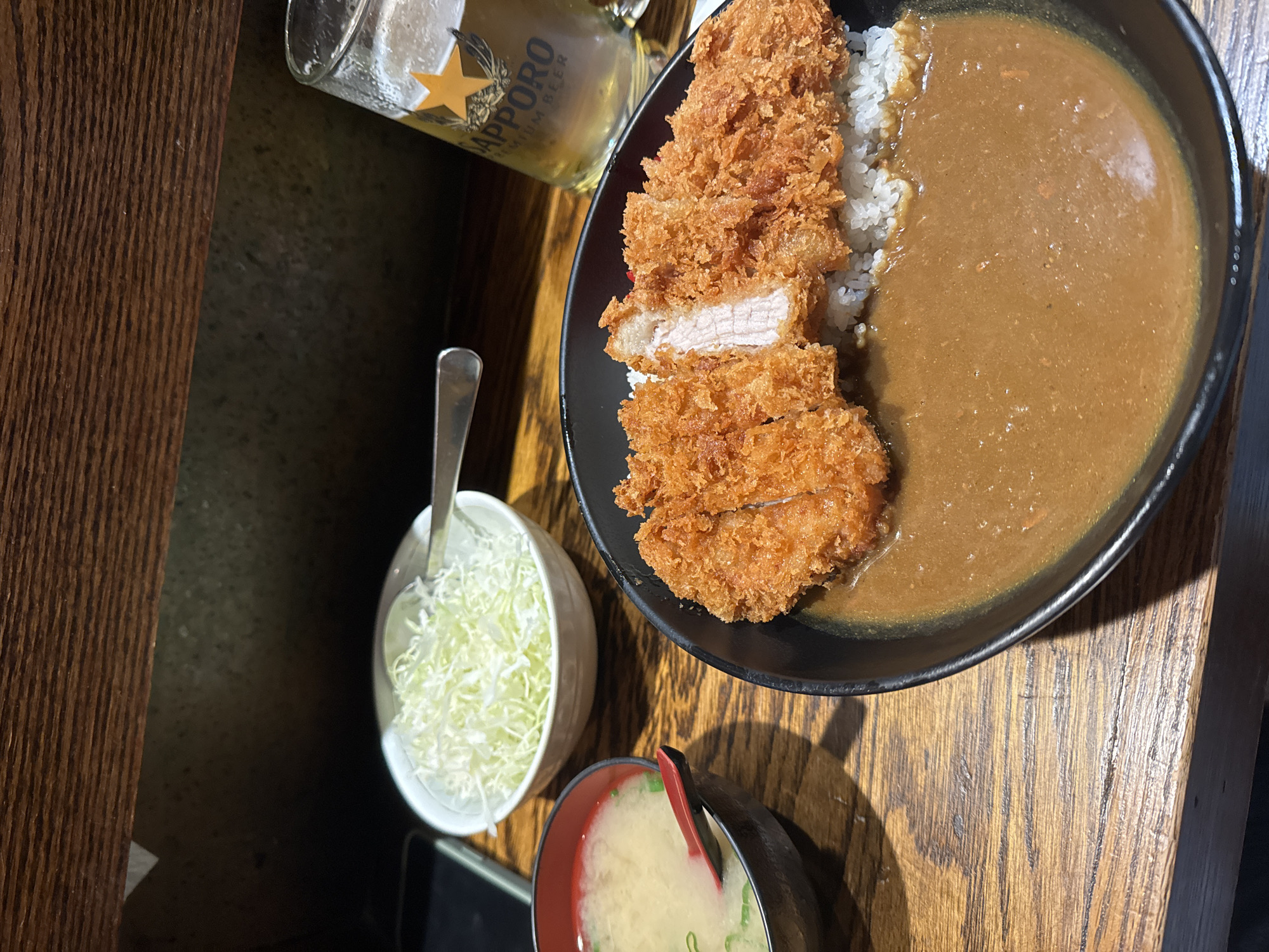 A meal is presented from an overhead perspective on a dark wooden table. The main dish, centered in a black bowl, consists of white rice covered by a substantial portion of sliced, fried tonkatsu (breaded pork cutlet) and brown Japanese curry sauce. One tonkatsu slice shows the pork meat. A smaller, black and red bowl to the left contains miso soup with green onion garnish and a red spoon. A white bowl holds finely shredded white cabbage, accompanied by a silver spoon. A clear glass mug, approximately half-full of amber liquid and foam, displaying the "SAPPORO PREMIUM BEER" logo and a gold star, is positioned on the right. A dark-colored smartphone is partially visible on the far left. The background features dark, textured wooden surfaces, indicative of a commercial dining establishment. The setting is identified as a Go! Go! Curry! restaurant located in New York City, New York, United States of America, based on the specific culinary presentation and restaurant branding elements.
