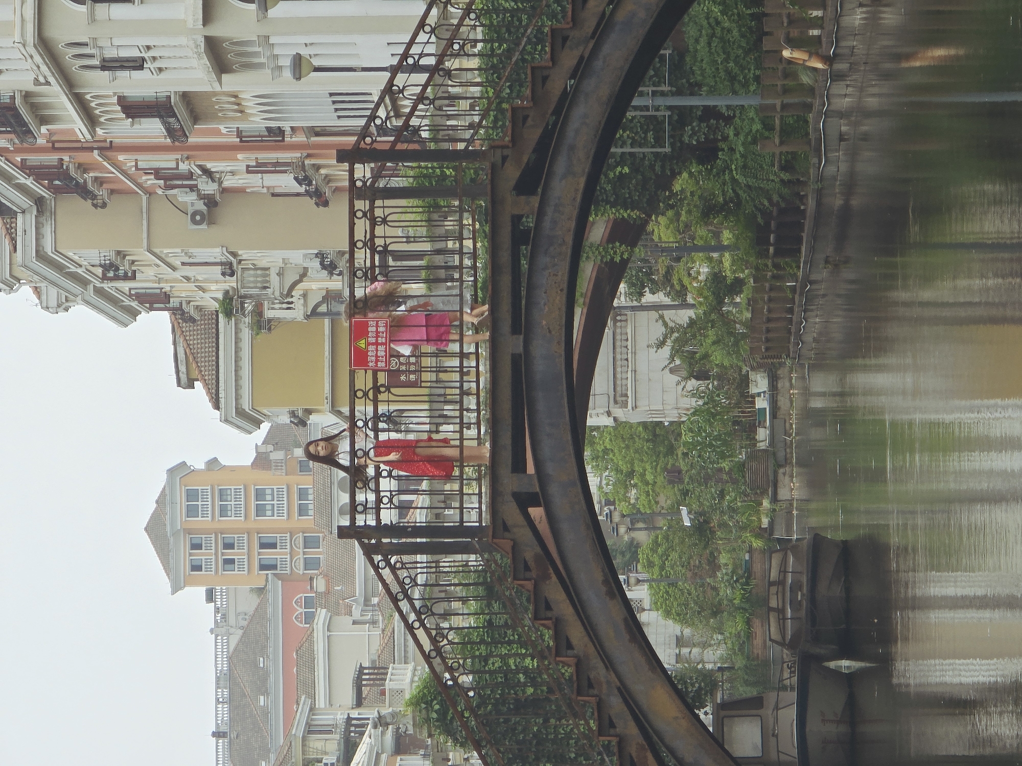 This is a medium shot of a canal in the Xiaoshan District of China. Two young females are standing on a rusty metal bridge that spans the canal. The female on the left is wearing a white top and a red skirt. The female on the right is wearing a pink dress. Both females are looking towards the camera. In the background, there are several multi-story buildings with European-style architecture. The buildings are painted in various colors, including yellow, orange, and white. There are trees and greenery on the sides of the canal. Two boats are docked on the left side of the canal. The sky is overcast.