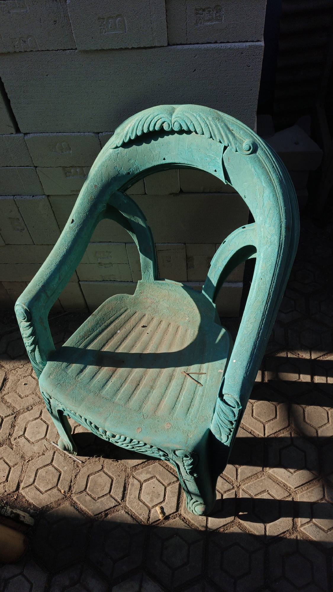 An aged, teal-colored plastic chair, possibly ornamental, rests on a hexagonal patterned concrete paver surface. The chair exhibits signs of wear and weathering, with some dirt and debris visible on its seat. It is positioned in front of a stack of light-colored concrete blocks, some of which have embossed lettering. Sunlight casts sharp shadows across the scene, suggesting it is daytime. The overall environment appears to be an outdoor, possibly industrial or storage area, located in Santing, Indonesia. No people or animals are present.