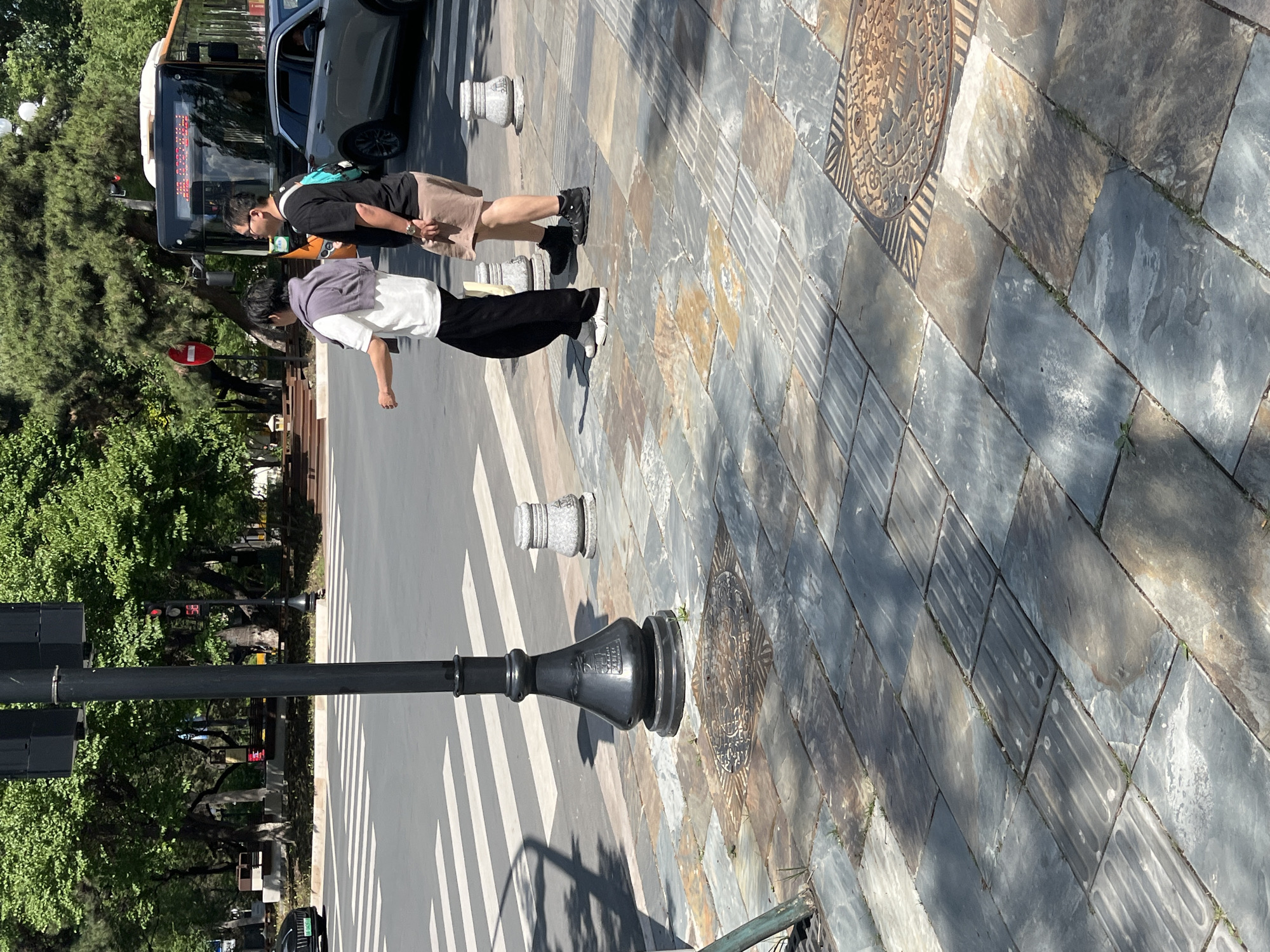 The image depicts a street scene in Changchun City, China. Two people are walking across a crosswalk. A bus is stopped nearby. A black lamppost is prominently featured in the foreground. The sidewalk is paved with patterned stones, including a visible manhole cover. Trees and a traffic light can be seen in the background. The sky appears sunny.