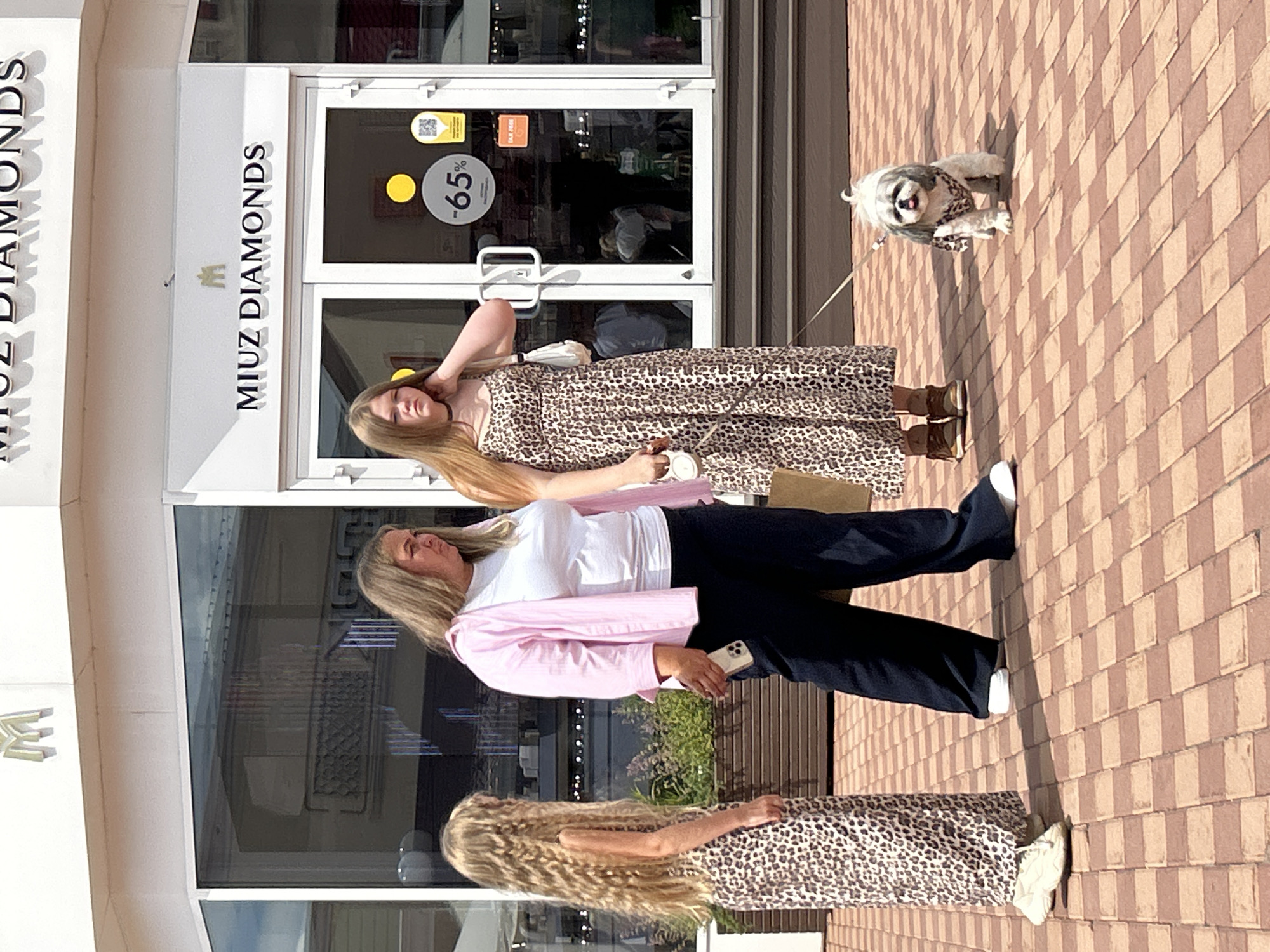 In the city of Pokrovskoe, Russia, three individuals and a small dog stand on a paved walkway in front of a storefront. The storefront, identified as "MIUZ DIAMONDS," features large glass windows and a white facade.
The person on the left, a young girl with long, wavy blonde hair, wears a leopard-print dress. To her right, a woman with blonde hair is dressed in a white t-shirt, a pink open shirt, and dark trousers. She is holding a smartphone in her right hand. The person on the far right, a young girl with straight blonde hair, is wearing a leopard-print dress and a black choker necklace. She is holding a leash attached to a small, white and grey dog. The dog is wearing a leopard-print bandana and is looking towards the camera with its tongue slightly out.
The ground is paved with reddish-brown rectangular tiles. In the background, through the shop windows, the interior of the store is visible, with displays and other shoppers. A sign on the window indicates a "-65%" discount. The overall scene suggests a sunny day.
