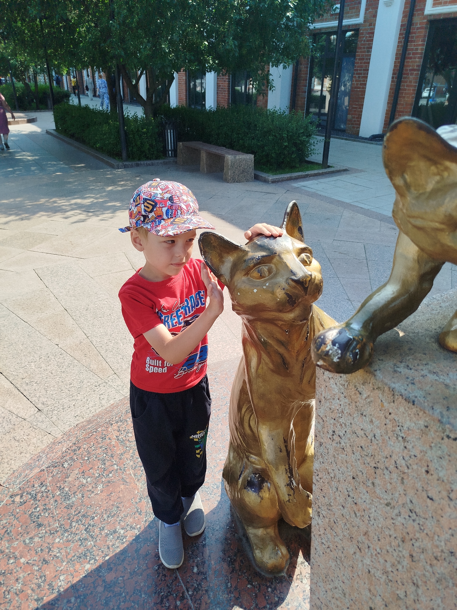 In the city of Tyumen, Russia, a young boy is pictured standing next to a golden cat statue. The boy, wearing a red t-shirt, black pants, and a colorful baseball cap, is touching the statue's head with his right hand. The statue is a bronze-colored sculpture of a cat, positioned on a stone base. The background reveals a public outdoor area with trees, bushes, a walkway, and buildings with brick accents. The lighting suggests it is daytime, with shadows cast on the ground.