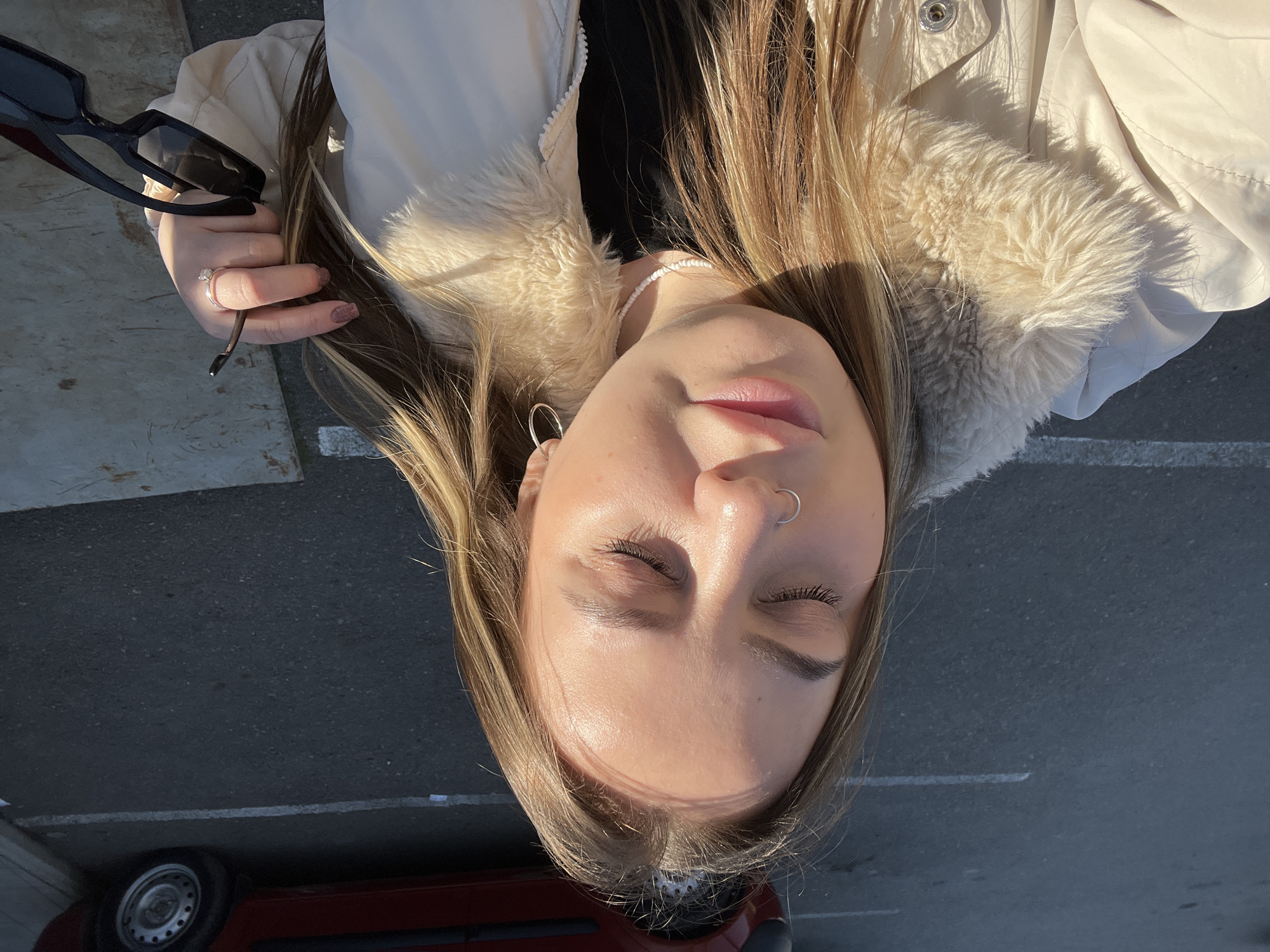 A woman with long blonde hair, closed eyes, and a nose ring is lying on a gray asphalt surface. She is wearing a light-colored jacket with a fur-lined collar and a white beaded necklace. Her left hand holds a pair of black sunglasses. The surface appears to be a parking lot, with a glimpse of a red vehicle's tire visible in the bottom left corner. The setting is outdoors during the day, with strong sunlight casting shadows. The location is indicated as the city of Novopokrovskaya, Russia.