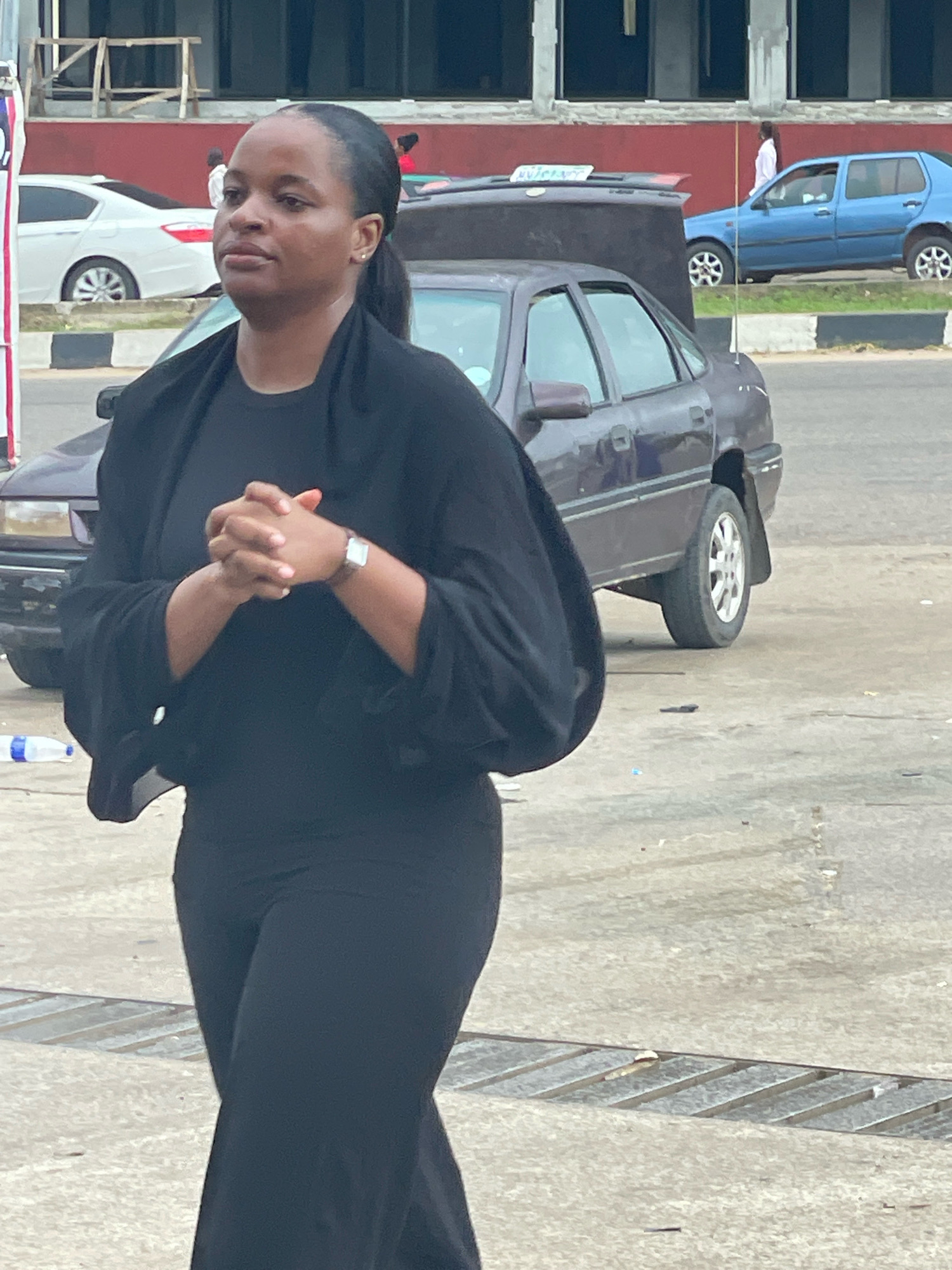 In a scene set outdoors on a paved surface, possibly a street or parking area, a Black woman is captured from the chest up, facing left. She wears a black garment with full, flowing sleeves and her hands are clasped in front of her chest. A silver-toned watch with a leather band is visible on her left wrist. Her hair is pulled back in a ponytail. In the background, a dark-colored car is parked, with its hood open. Further behind, a light blue sedan is visible, and to the left, a white sedan. The environment suggests an urban setting in Abuja, Nigeria, with a glimpse of building structures and what appears to be a drainage grate on the ground in the foreground.