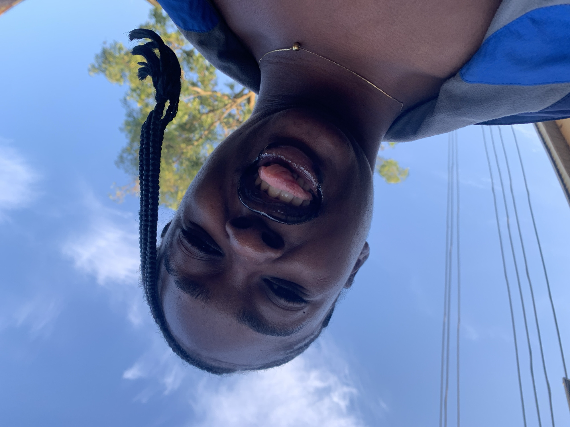 An eye-level, wide shot captures a young Black person from the chest up, looking up at the camera. The person's head is tilted back, and their mouth is open with their tongue sticking out. They have dark hair styled in braids, with one braid extending over their head and towards the top right of the frame. The person is wearing a gold chain necklace with a small ball pendant. They are also wearing a gray and blue jacket. The background consists of a bright blue sky with scattered white clouds, a leafy green tree, and power lines. The image appears to be taken outdoors. It is not possible to determine the specific location within Rafin Par, Nigeria from this image.
