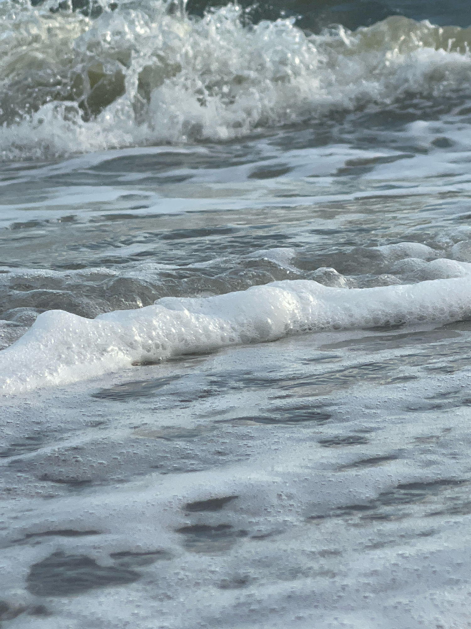 The image depicts a close-up view of ocean waves and foam at Pattaya City, Thailand. In the background, a wave is breaking, creating a frothy white crest. In the foreground, a line of white foam created by receding waves stretches across the frame. The water itself is a mix of gray and blue tones, with subtle ripples and reflections visible. The lighting suggests it is daytime, with sunlight glinting off the water's surface. There are no people or man-made objects visible in the image.