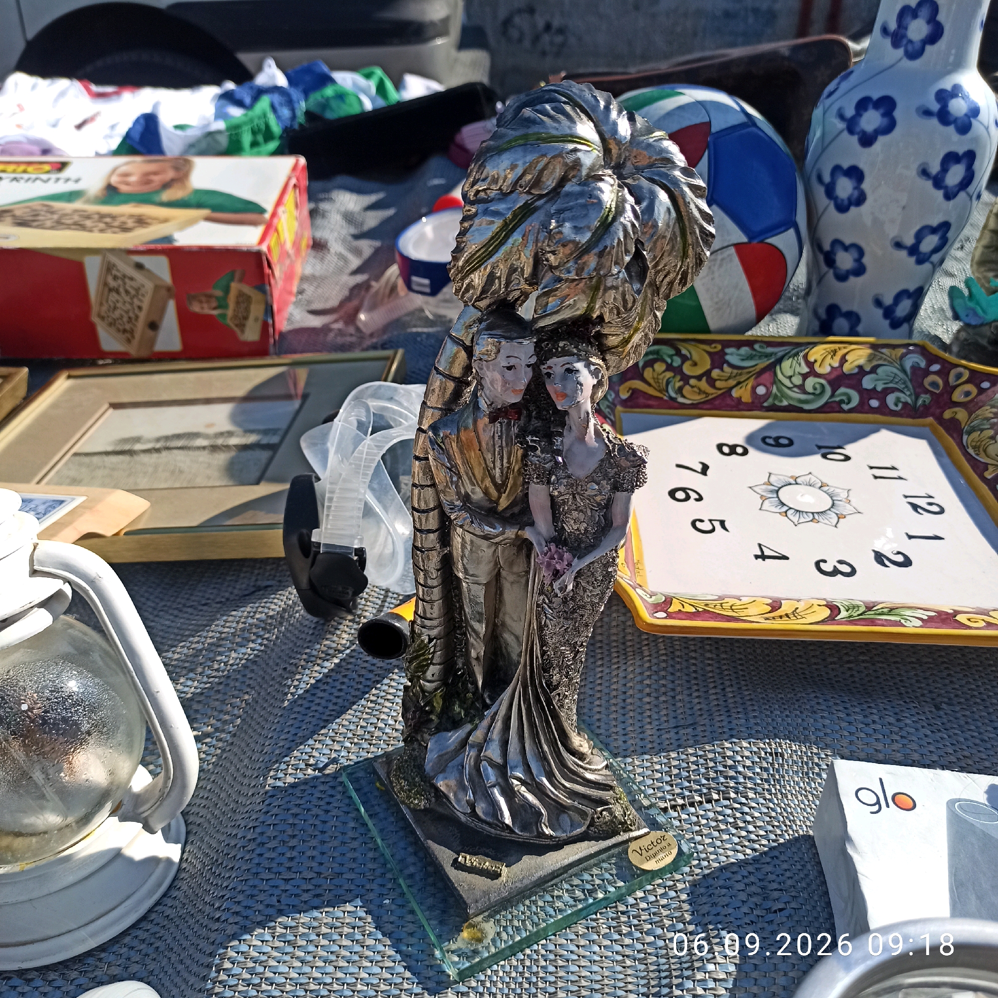 The image depicts an outdoor market scene, likely a flea market, set up on a woven blue and grey mat. The main subject is a silver-colored figurine of a bride and groom, presented in a formal attire, standing close together. The groom wears a suit with a bowtie, and the bride is adorned in a detailed dress, holding a small bouquet. They are positioned on a rectangular glass base, which has an inscription on it.
Scattered around the figurine are various items. To the left, a white kerosene lantern with a glass globe is visible, showing reflections within. Behind the figurine, to the left, is a red cardboard box with a picture of a child and the word "LABYRINTH" printed on it. To the right of the figurine, there is a decorative plate with a clock face and colorful floral patterns on its border. Further to the right, a blue and white patterned vase is present. In the background, other market items and fabrics, including a multi-colored ball, are visible.
The lighting suggests it is daytime, with shadows cast by the objects. The scene is cluttered, typical of a market stall. The timestamp at the bottom right of the image reads "06.09.2026 09:18", indicating the date and time the photo was taken. The question of location in Sannicandro di Bari, Italy, cannot be determined from the visual information in the image.