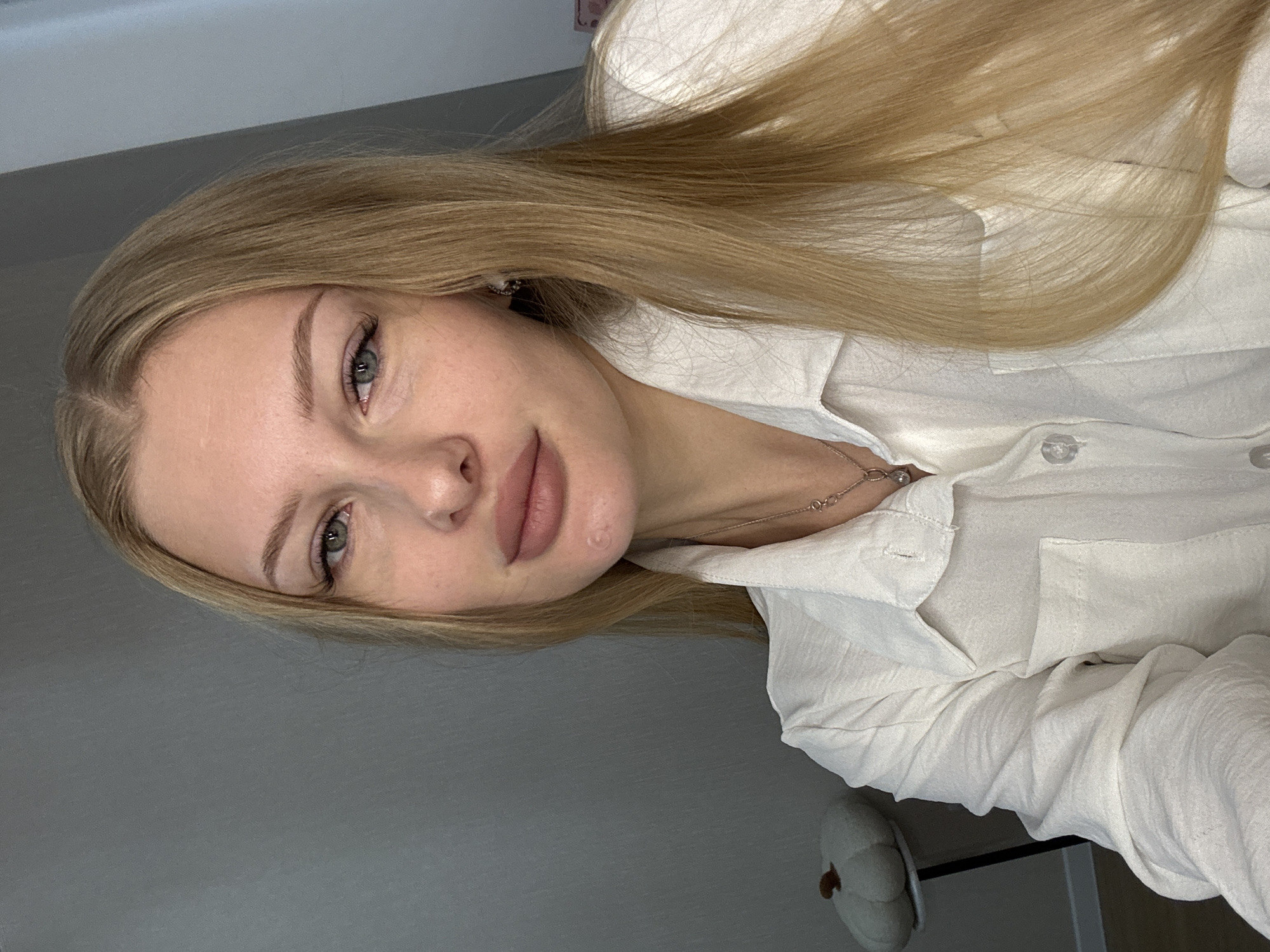 The image shows a close-up, medium shot of a young Caucasian woman with long blonde hair, fair skin, and light-colored eyes. She is wearing a white collared shirt. Her lips are slightly parted, and she is looking towards the camera. The background is a plain gray wall, with a white decorative pumpkin on a stand to the left of the woman. There is no discernible location in the city of Perm, Russia.