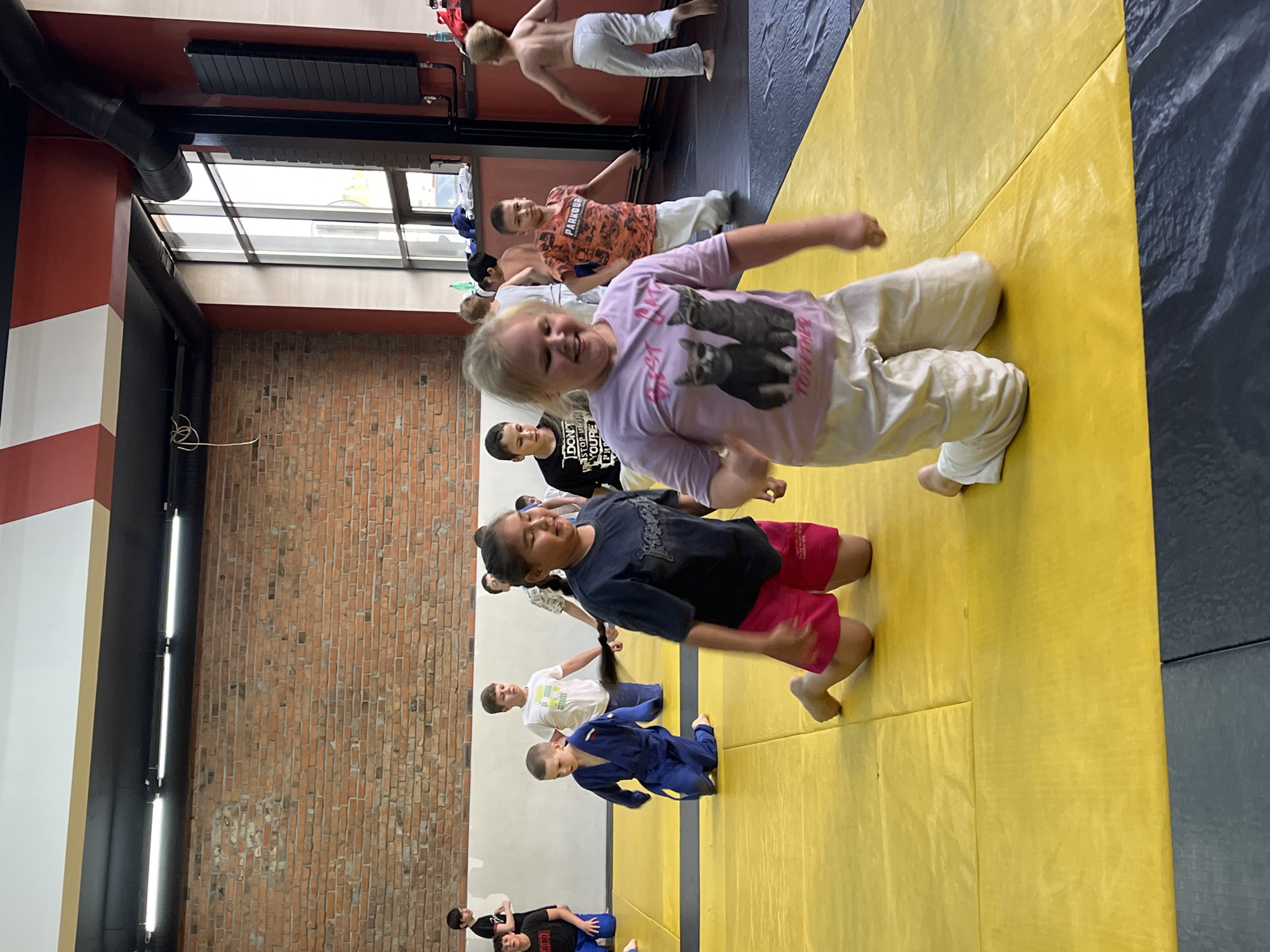 The image depicts an indoor martial arts training session, likely a dojo, with young participants. The environment features a yellow mat surface, a brick wall, and large windows. In the foreground, a blonde girl in a light purple t-shirt with a cat graphic and beige pants is on her knees, smiling and looking towards the viewer. To her left, a girl with dark hair in a dark t-shirt and pink shorts is also on her knees. Several other children are visible in the midground and background, some wearing martial arts uniforms and others in casual athletic wear. One boy in the midground is wearing a blue uniform. The overall scene suggests a group activity or lesson. The specific location in Krasnoyarsk, Russia cannot be determined from the image alone.