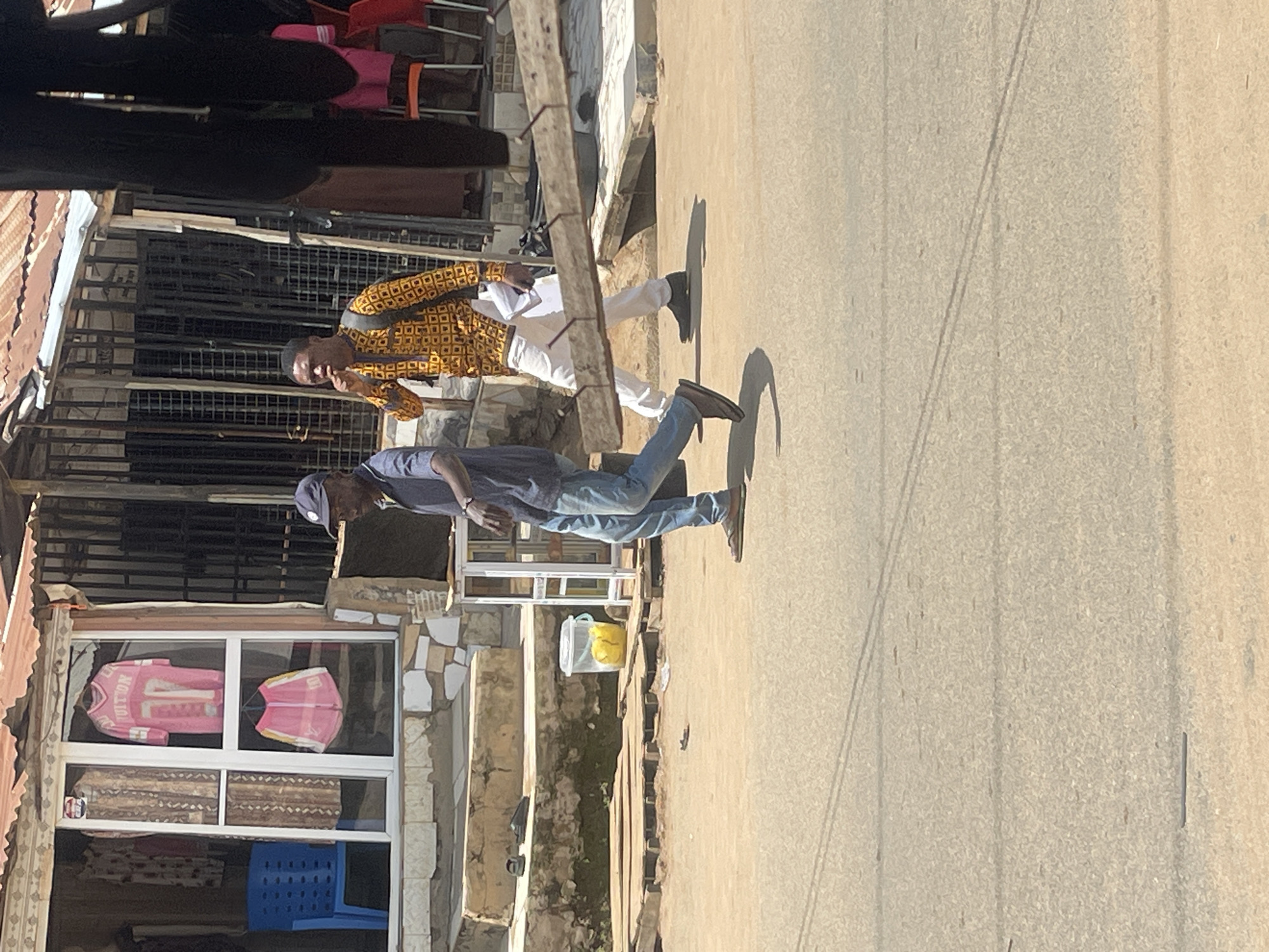 The image depicts a street scene in Abuja, Nigeria. In the foreground, two men are walking on a paved road. The man on the left, wearing a blue shirt, blue jeans, and a baseball cap, is walking towards the right side of the frame. The man on the right, dressed in a yellow patterned shirt and white trousers, is also walking towards the right, with a backpack on his shoulders and his hand near his mouth.
Behind the men, there is a building with a storefront. The storefront features a display of clothing, including pink garments with white lettering. A blue plastic chair is visible inside the store. The building has a corrugated metal roof and is situated on a street with a dirt shoulder. The environment appears to be an outdoor market or commercial area. A yellow bucket is placed on the ground near the building. The overall lighting suggests it is daytime.
